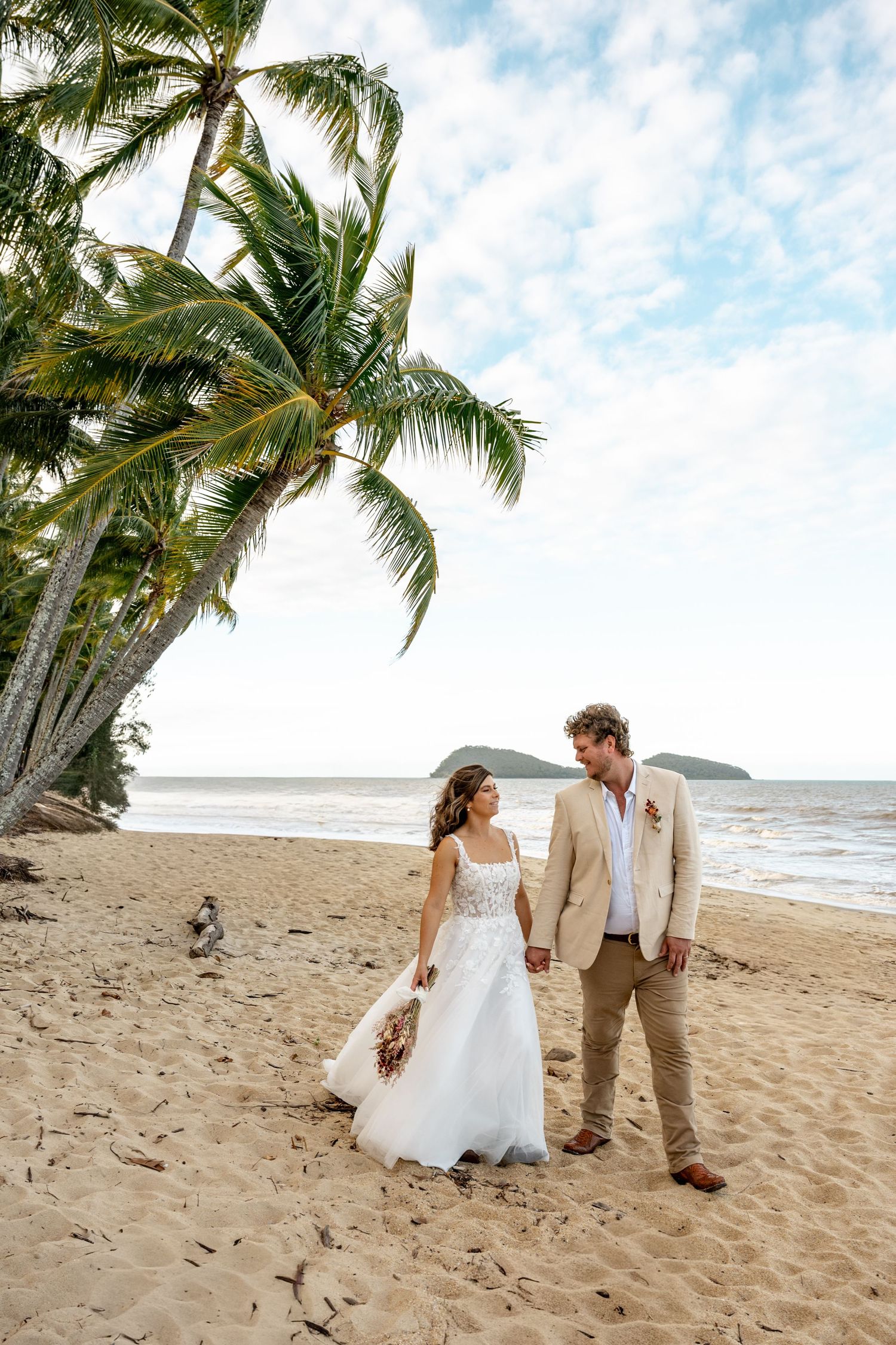 Newlyweds share a loving gaze while holding hands on a palm-fringed tropical beach with ocean backdrop.
