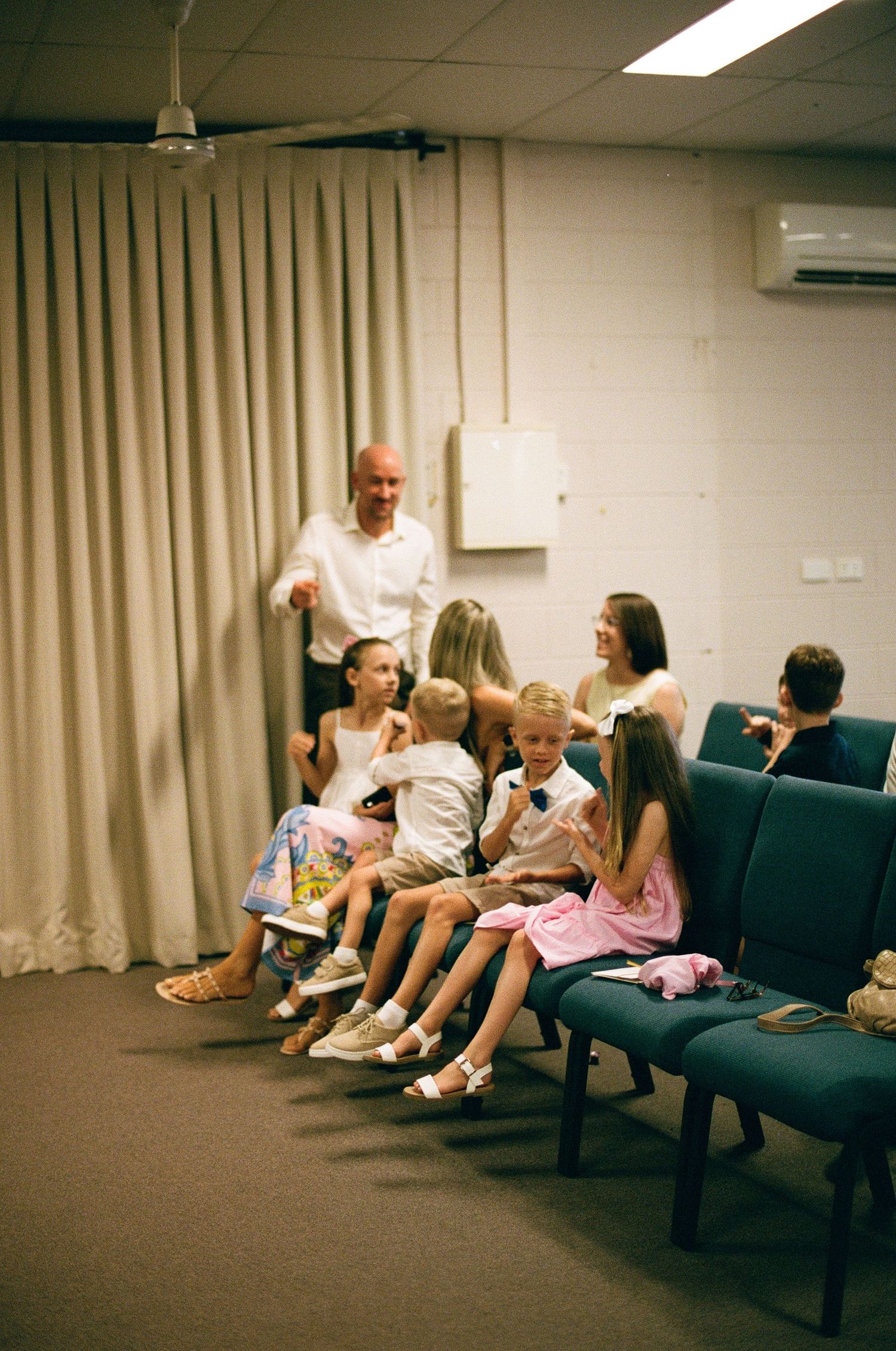 Children sitting on teal chairs at wedding venue while adult man gestures nearby.