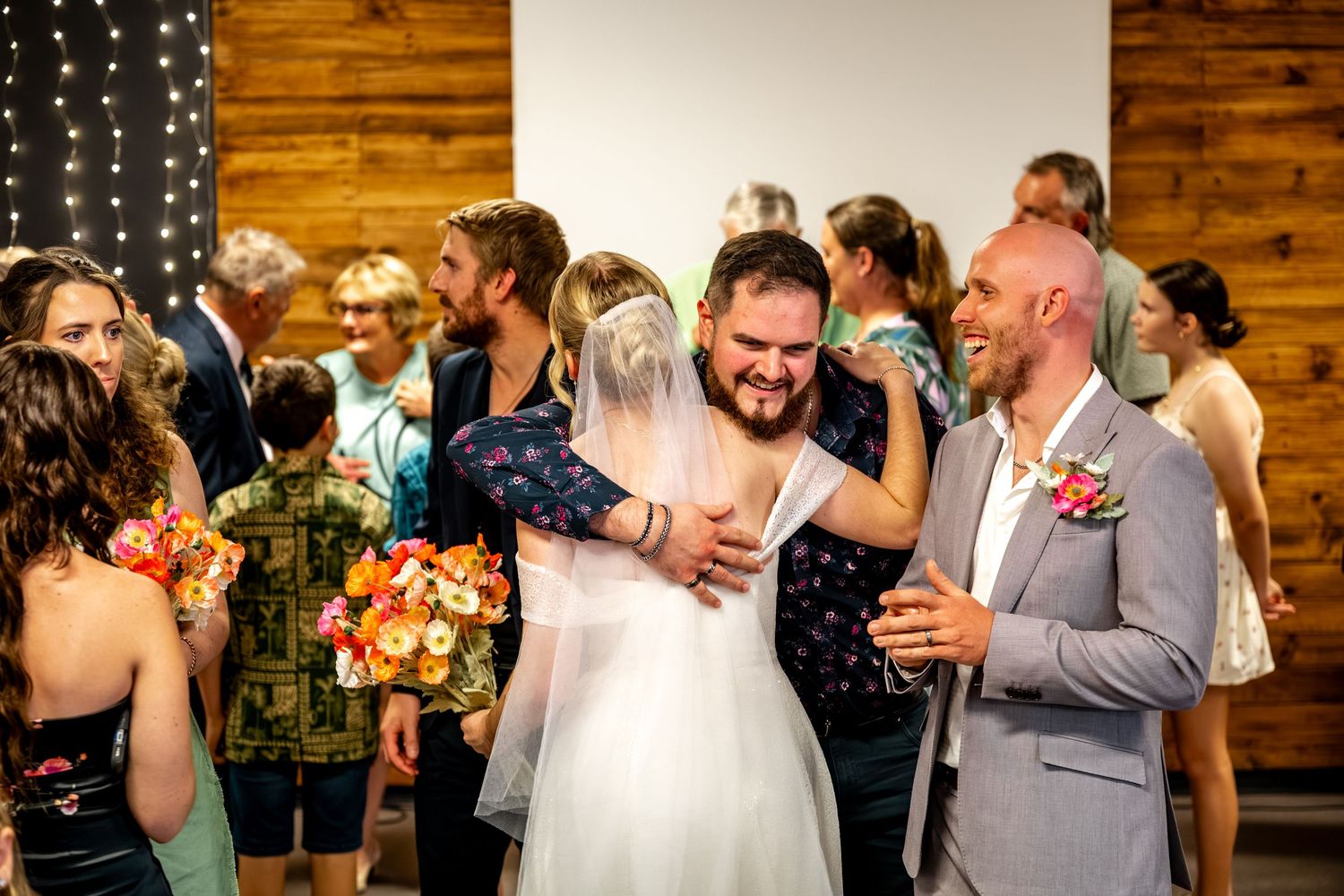 Groom in grey suit laughs as bride embraces guest during warm post-ceremony celebrations.