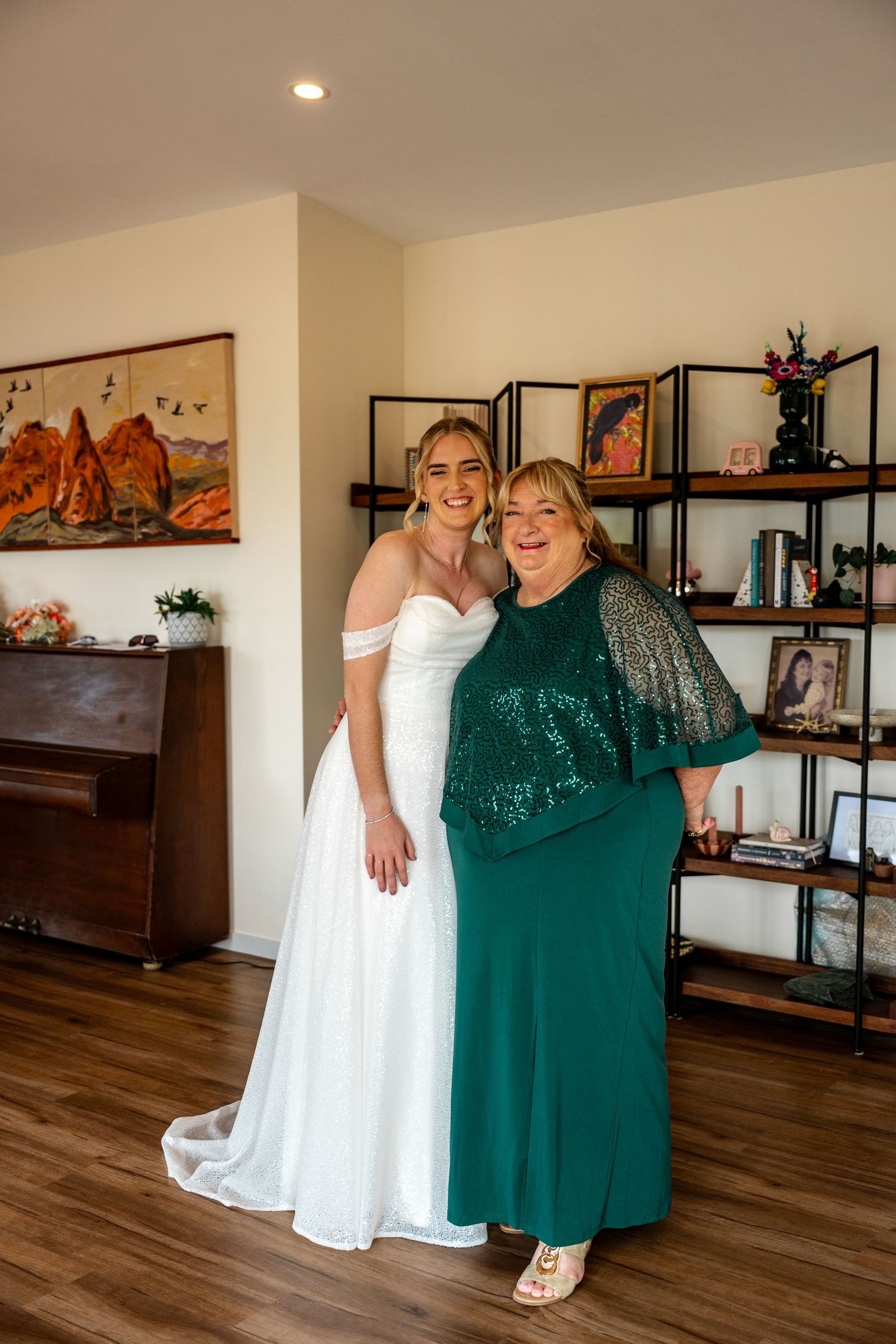 Bride in white off-shoulder gown smiling with mother in emerald green sequin dress.