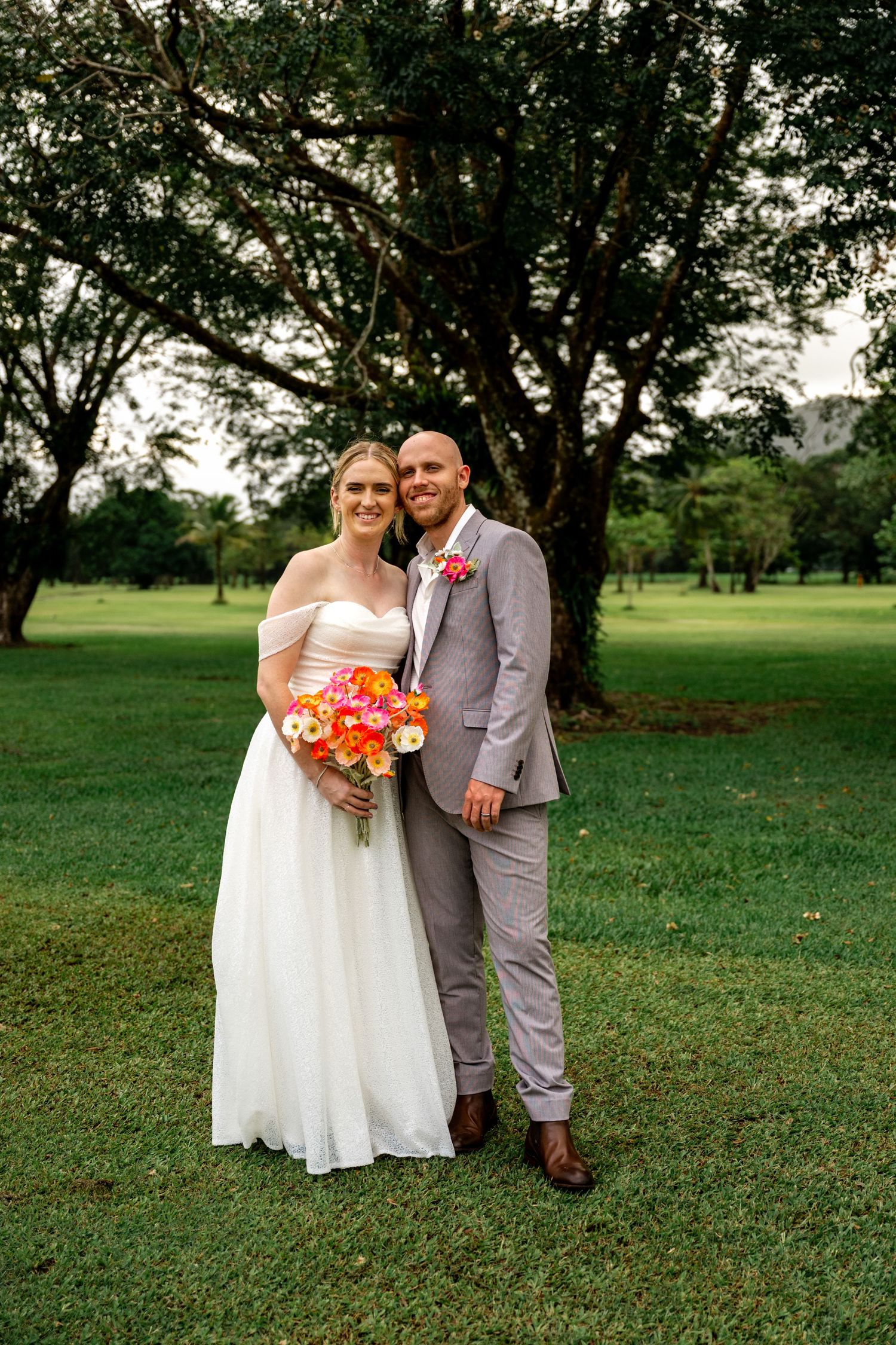 Wedding couple posing together on green lawn, bride in white off-shoulder gown holding colorful bouquet, groom in grey suit.