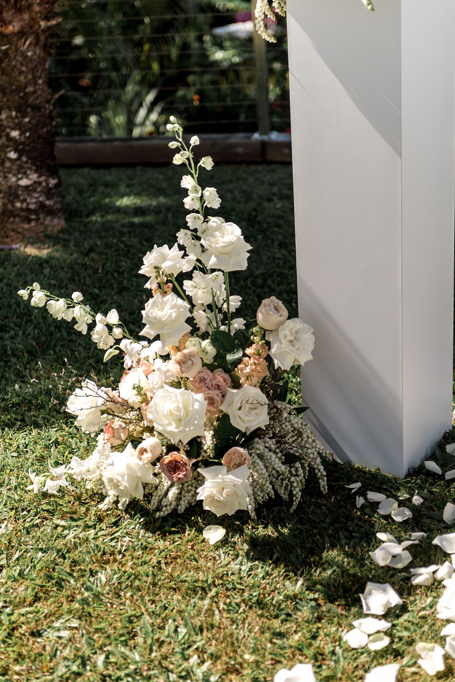 Cascading white and blush wedding flowers arranged at the base of a white ceremony pillar on a green lawn.