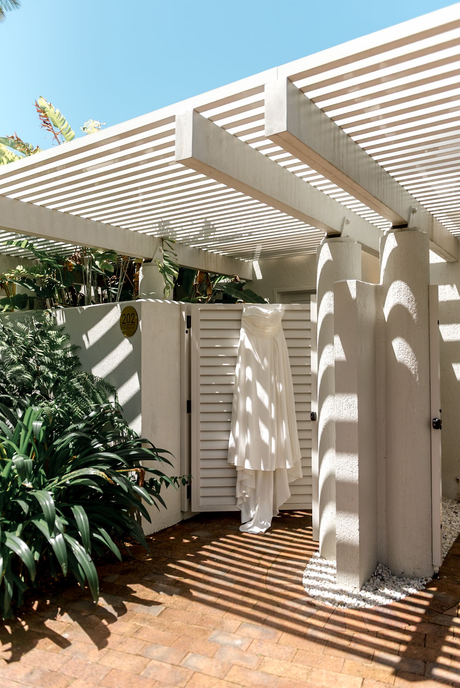 White pergola with louvered roof casting shadows over an outdoor shower area surrounded by tropical greenery.