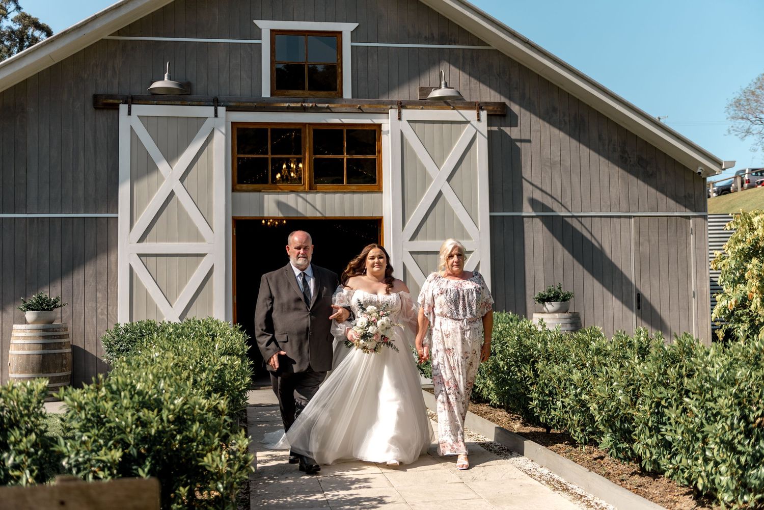 Bride in white gown walks with parents outside a charming gray barn wedding venue.