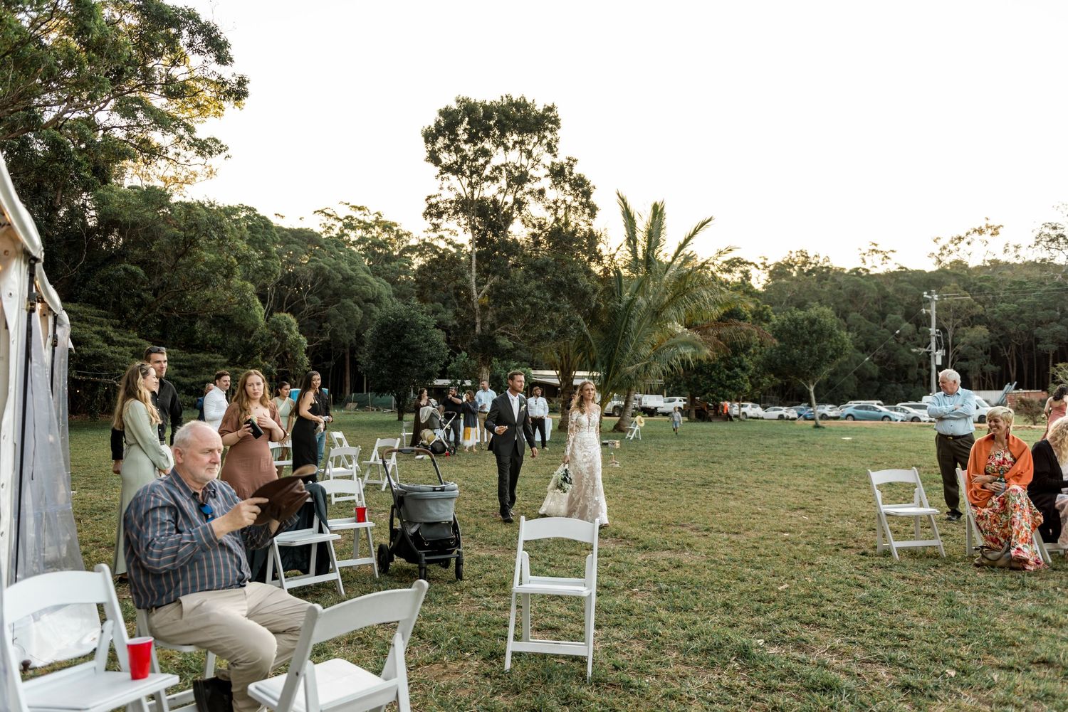Wedding guests seated on white chairs watch a ceremony on a green lawn surrounded by tall eucalyptus trees.