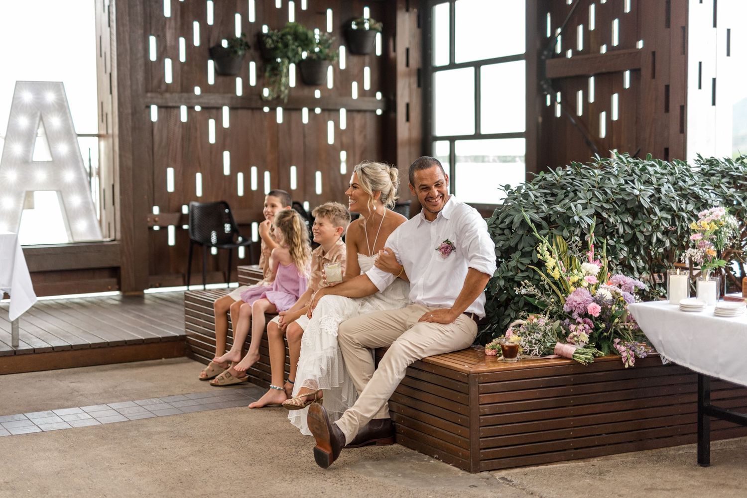 Bride and groom seated with children at a modern tropical wedding reception with wooden decor.
