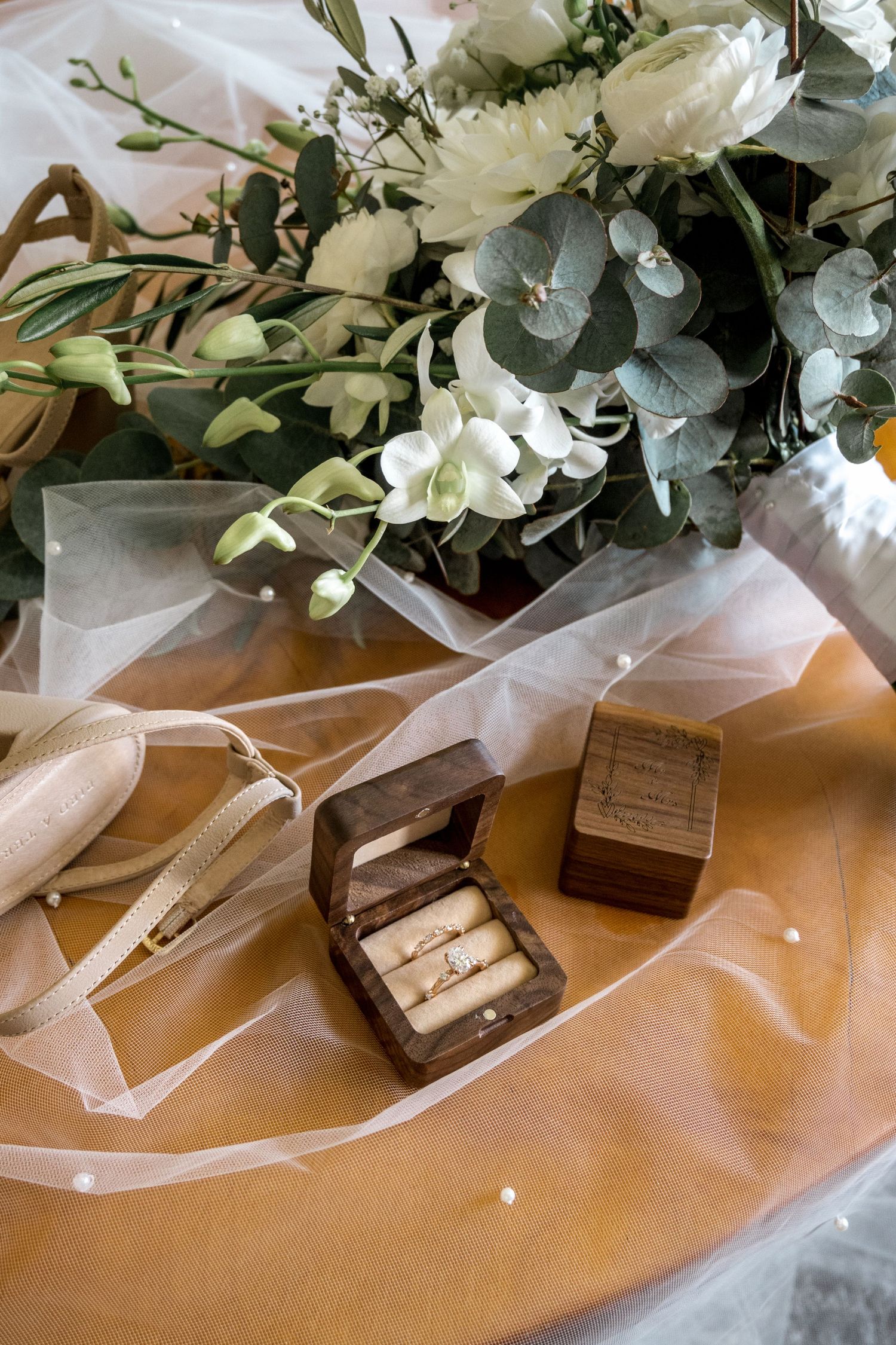 Elegant wedding flat lay featuring gold rings in wooden box, white floral bouquet, and sheer veil.