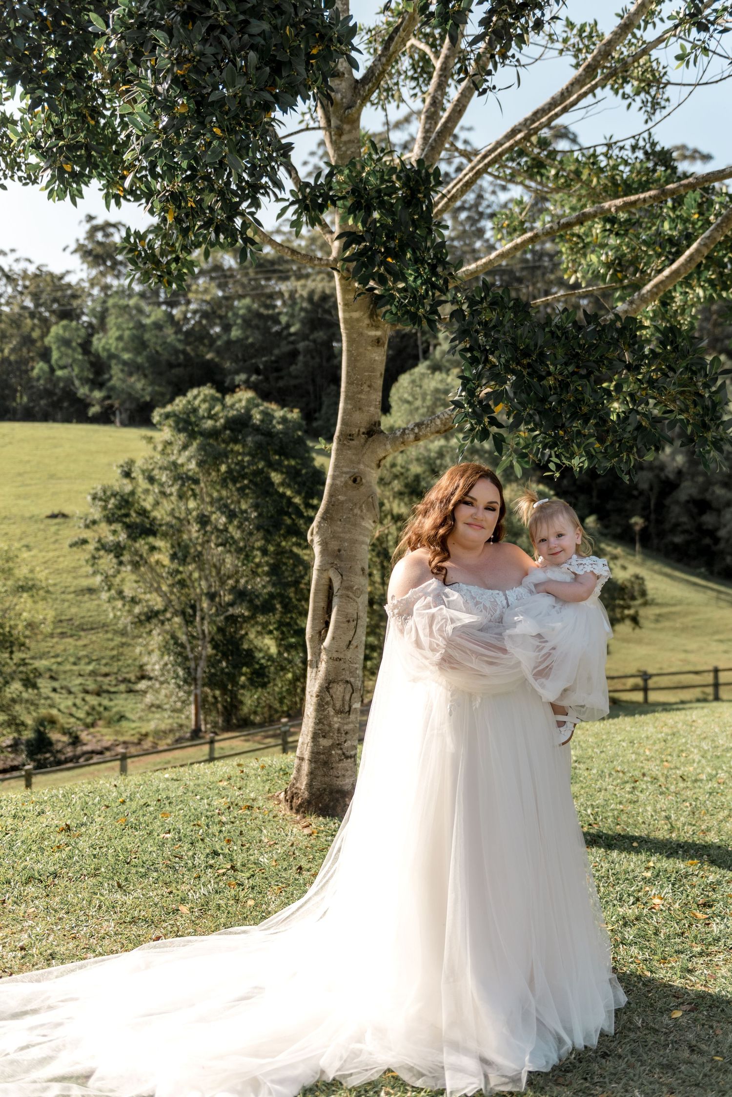 Bride in flowing white tulle gown holding baby in white outfit in lush green countryside setting.