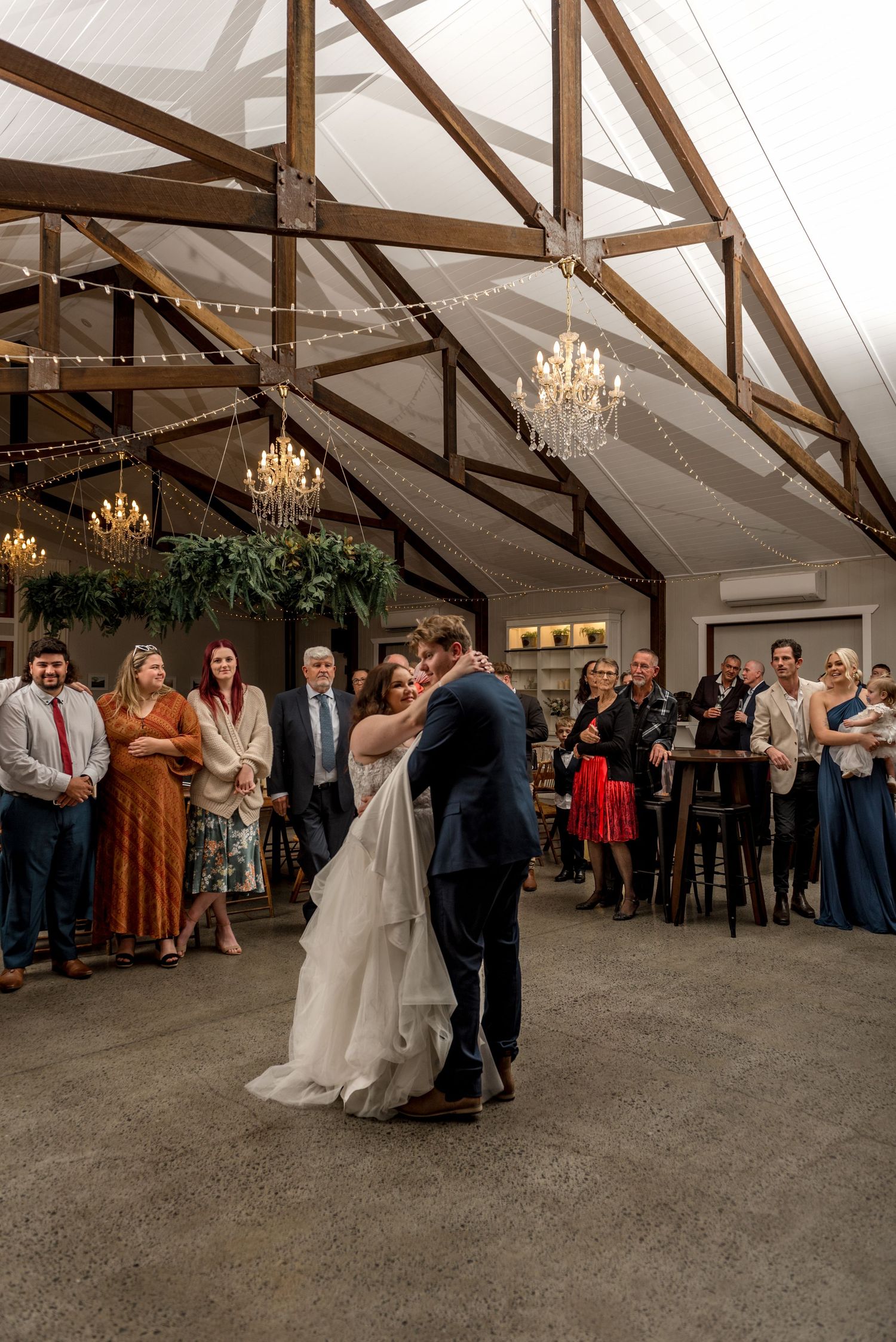 Bride and groom share their first dance in an elegant barn wedding venue with chandeliers and string lights.