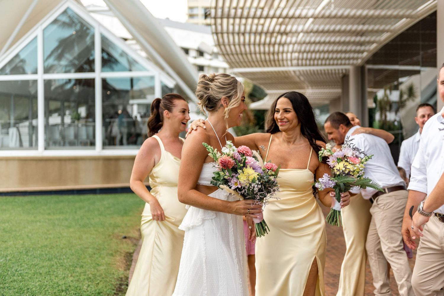 Bride walks with bridesmaids in yellow satin gowns holding colorful bouquets at a tropical venue.