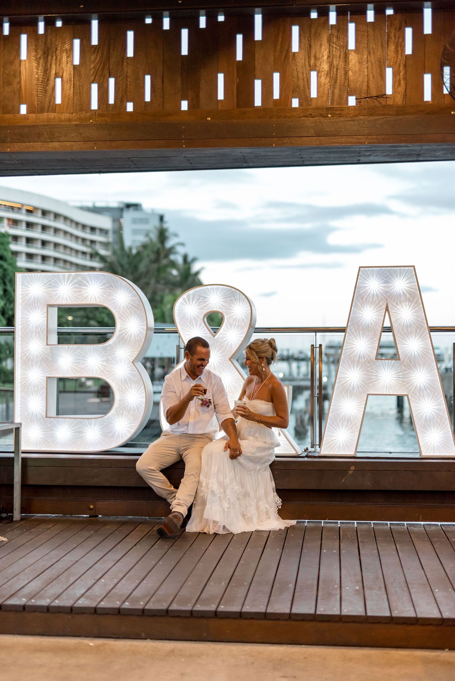 Bride and groom sitting together in front of illuminated B&A marquee letters at wedding reception.