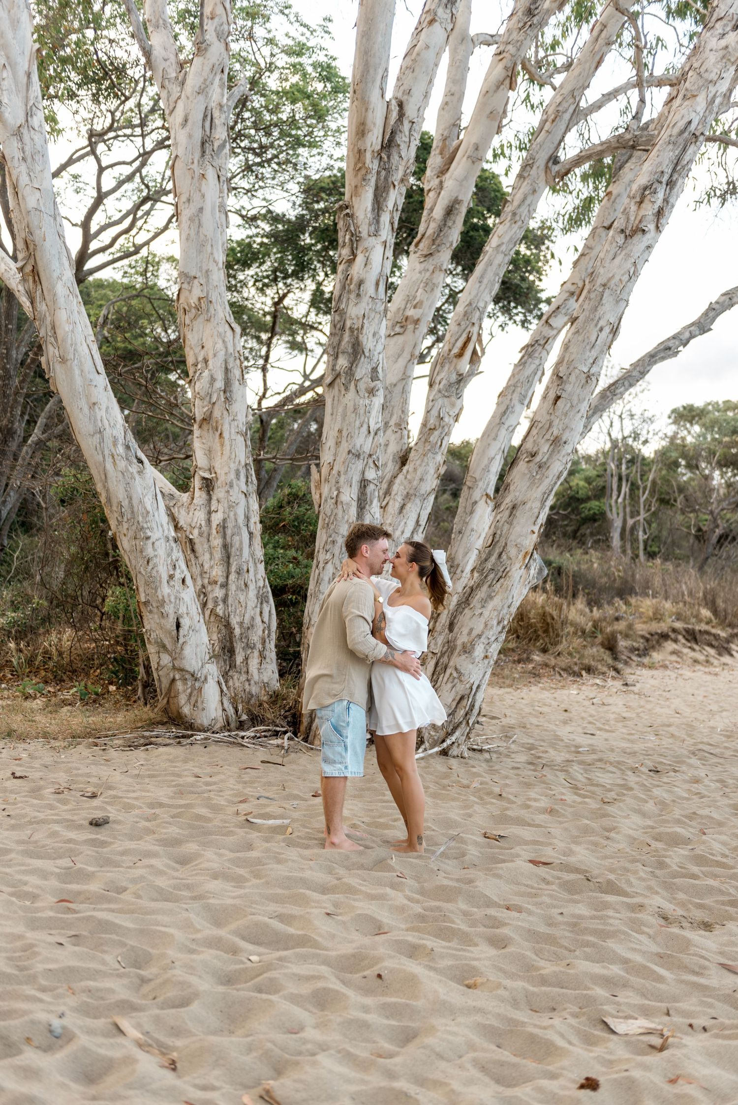 Couple sharing a kiss on sandy beach with large white paperbark trees in background.