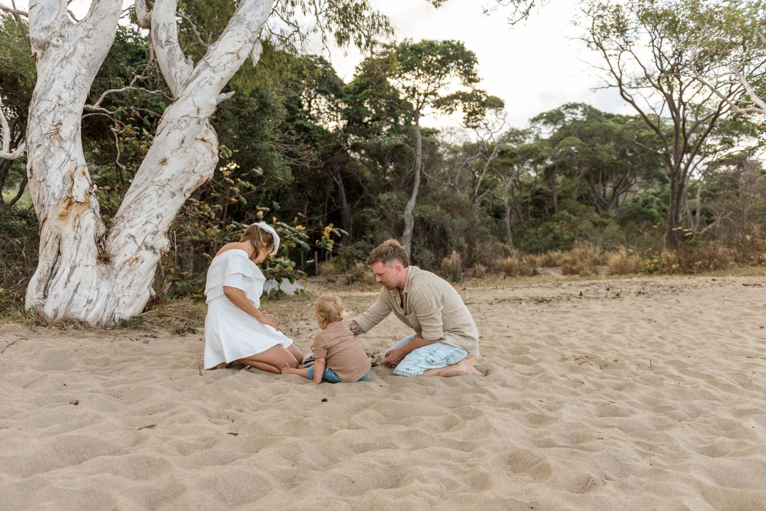 A young family sits together on sandy beach near white-trunked trees enjoying quality time.