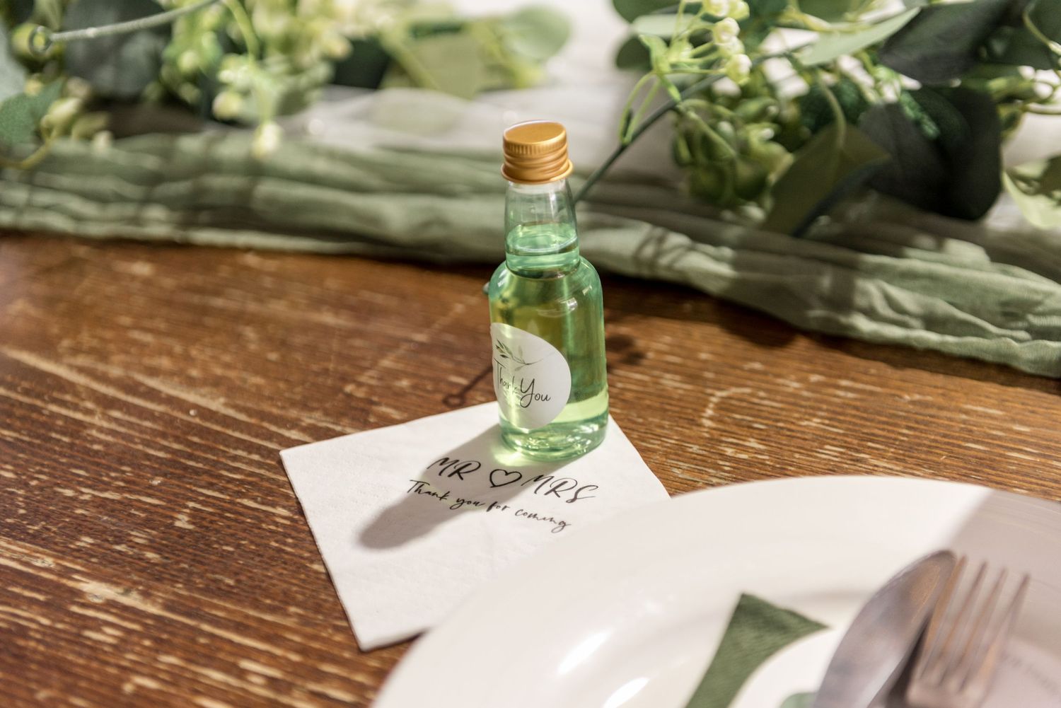 Mini green bottle wedding favor with personalized napkin on rustic wooden reception table.