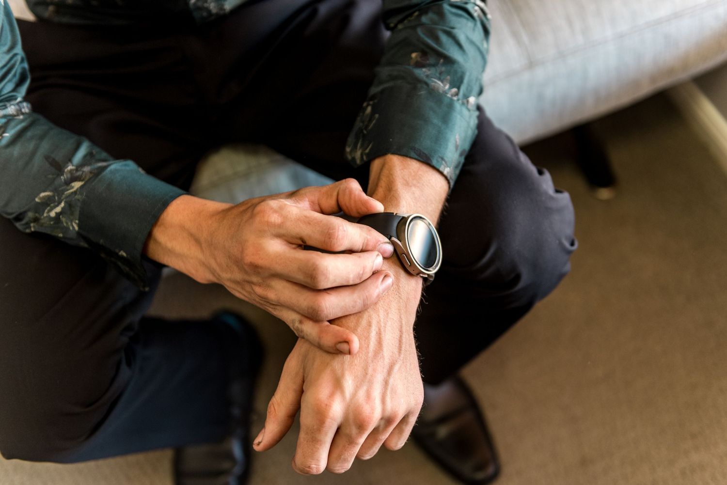 Close-up of a mans hands adjusting a black smartwatch while seated, wearing a dark patterned shirt.