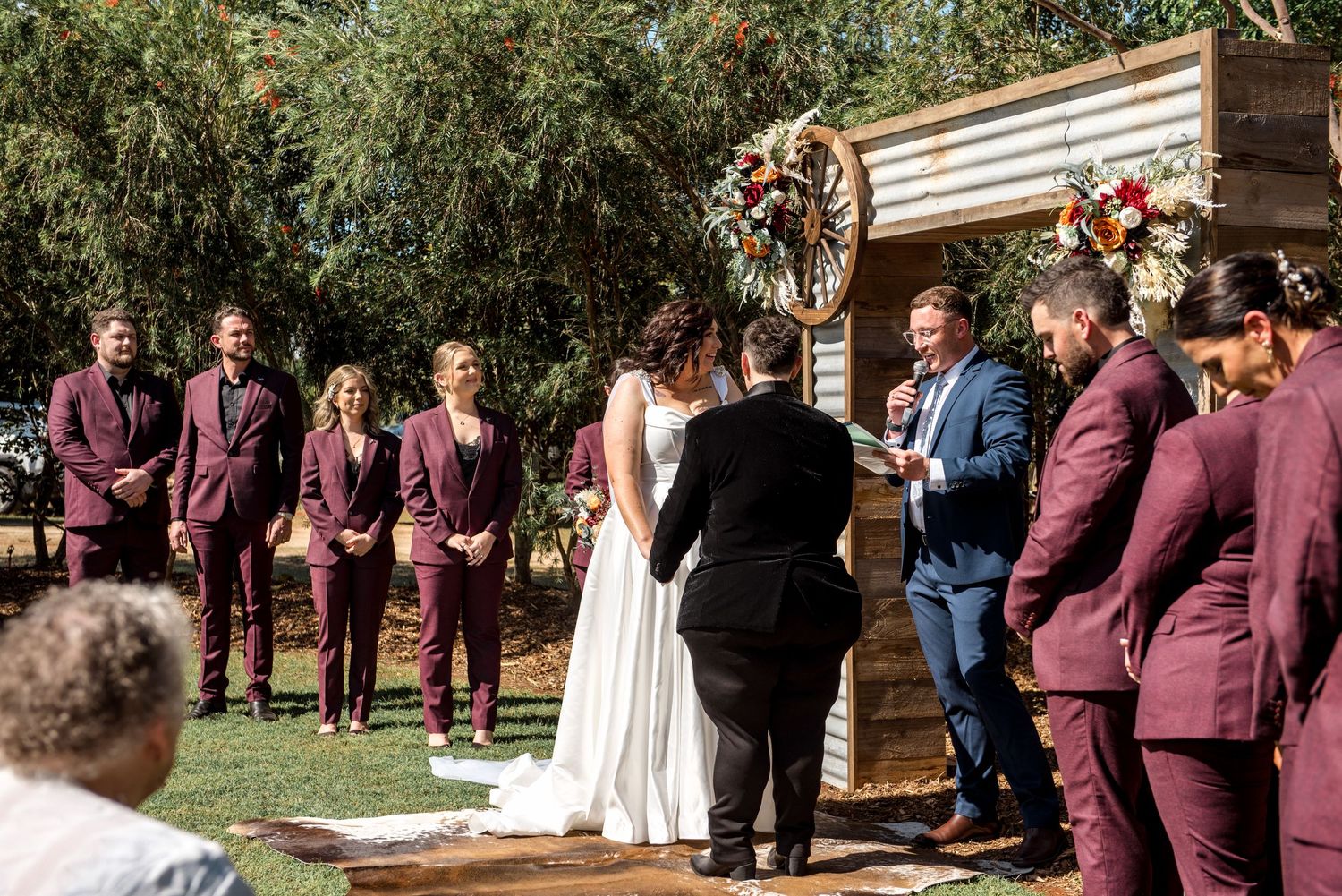 Outdoor wedding ceremony with bride and groom at rustic wooden altar, flanked by burgundy-suited groomsmen and bridesmaids.