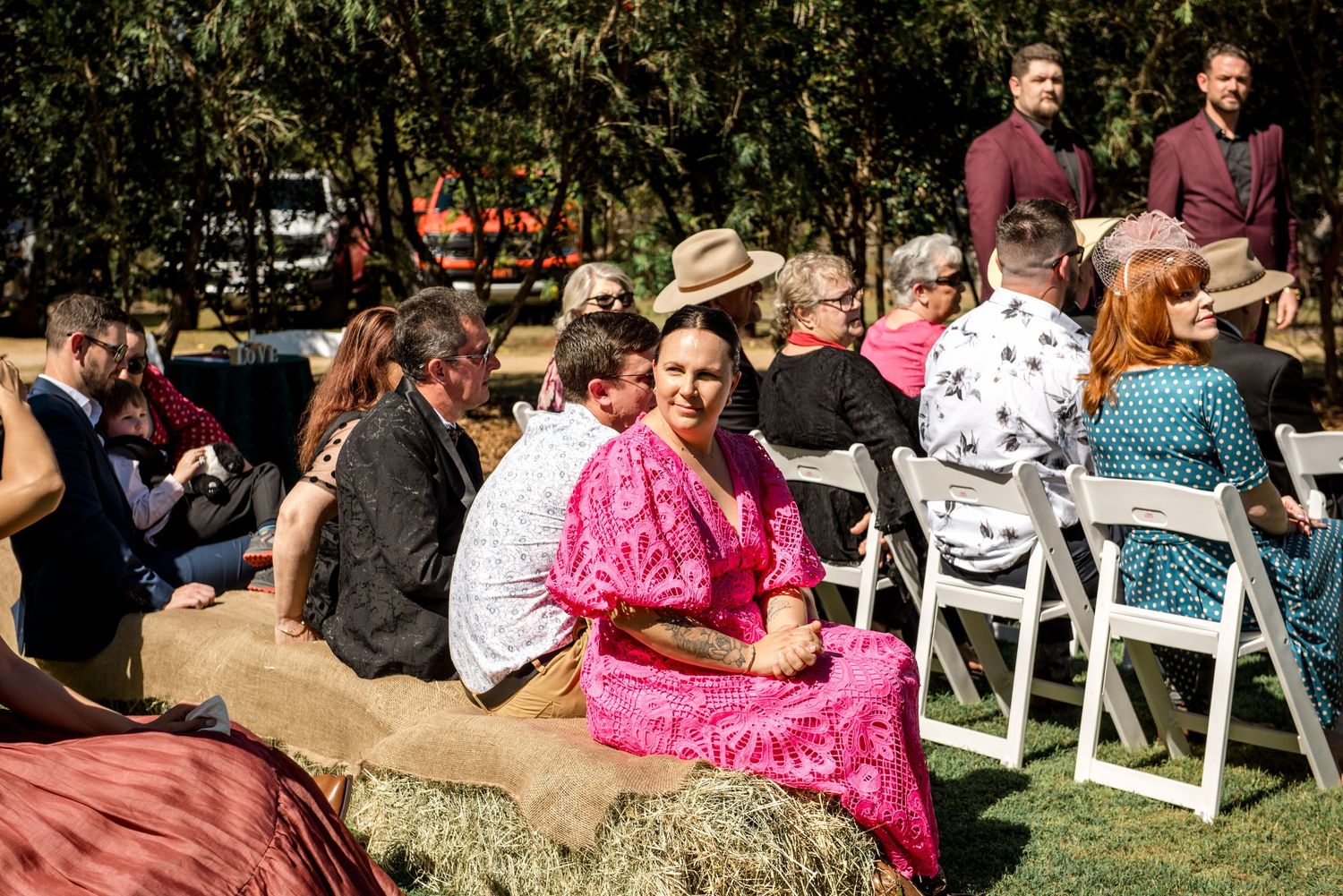 Smiling wedding guest in bright pink dress sits on hay bale seating at rustic outdoor ceremony.