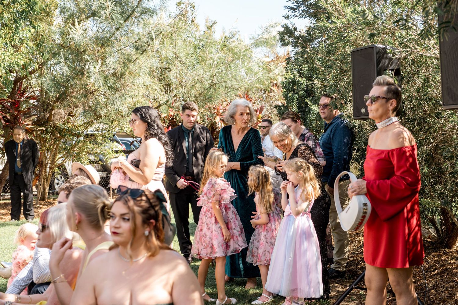 Wedding guests mingling outdoors among trees, including children in floral dresses and women in red and green.