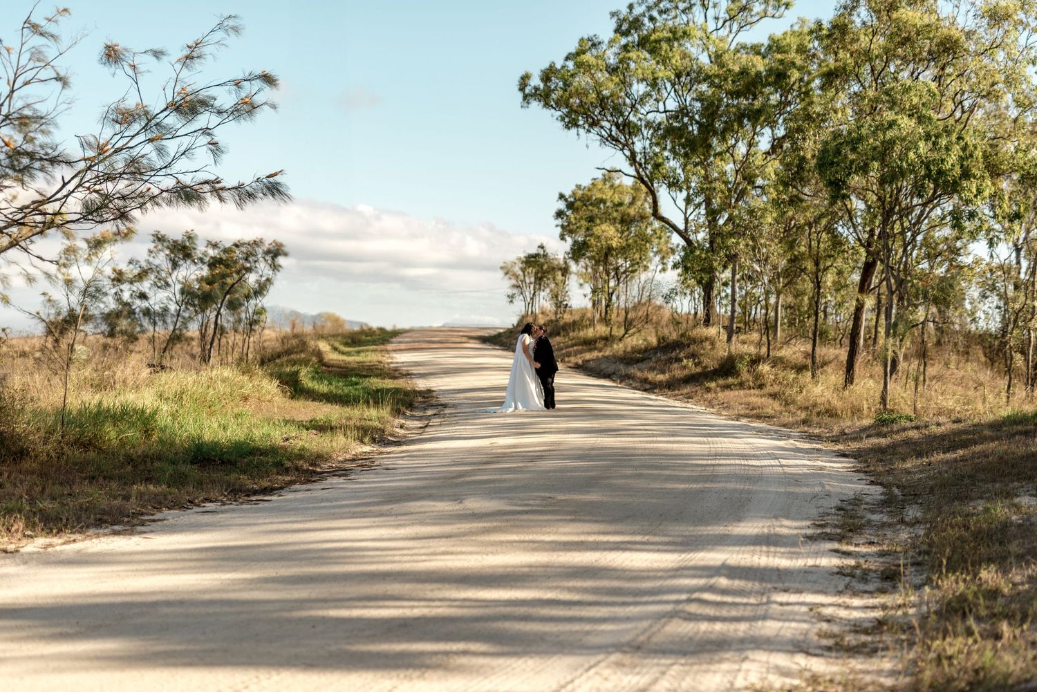 Same-sex newlyweds walking hand-in-hand along a sunlit straight road lined with Australian eucalyptus trees.