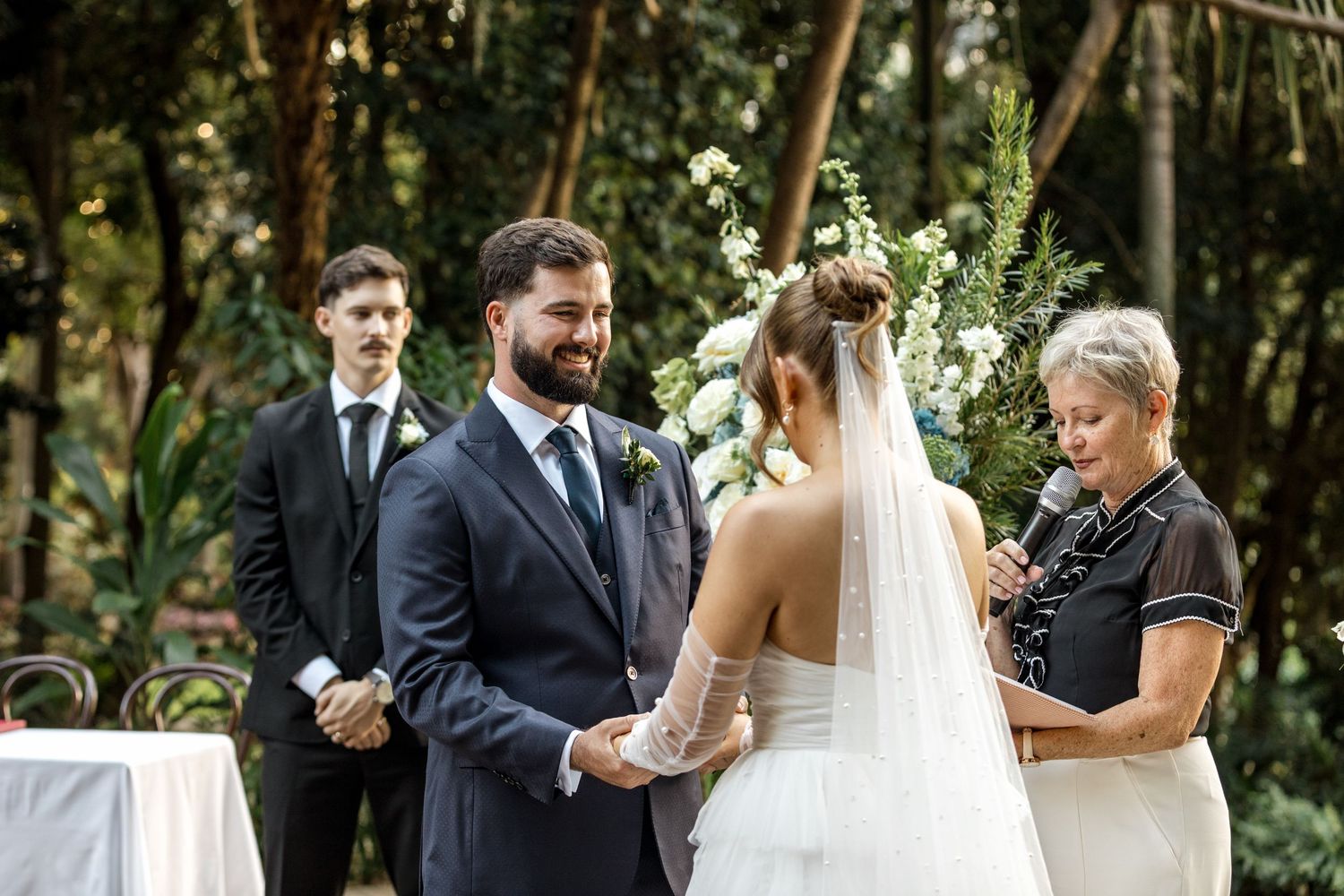 Smiling groom in navy suit holds hands with bride as officiant reads vows in garden ceremony.