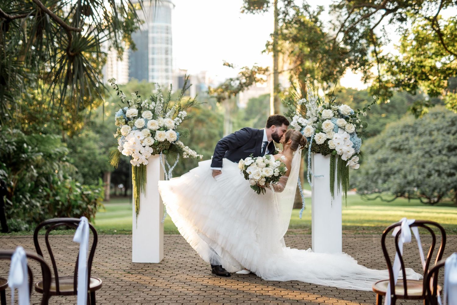 Groom dips bride for a kiss at outdoor wedding altar surrounded by white floral arrangements.