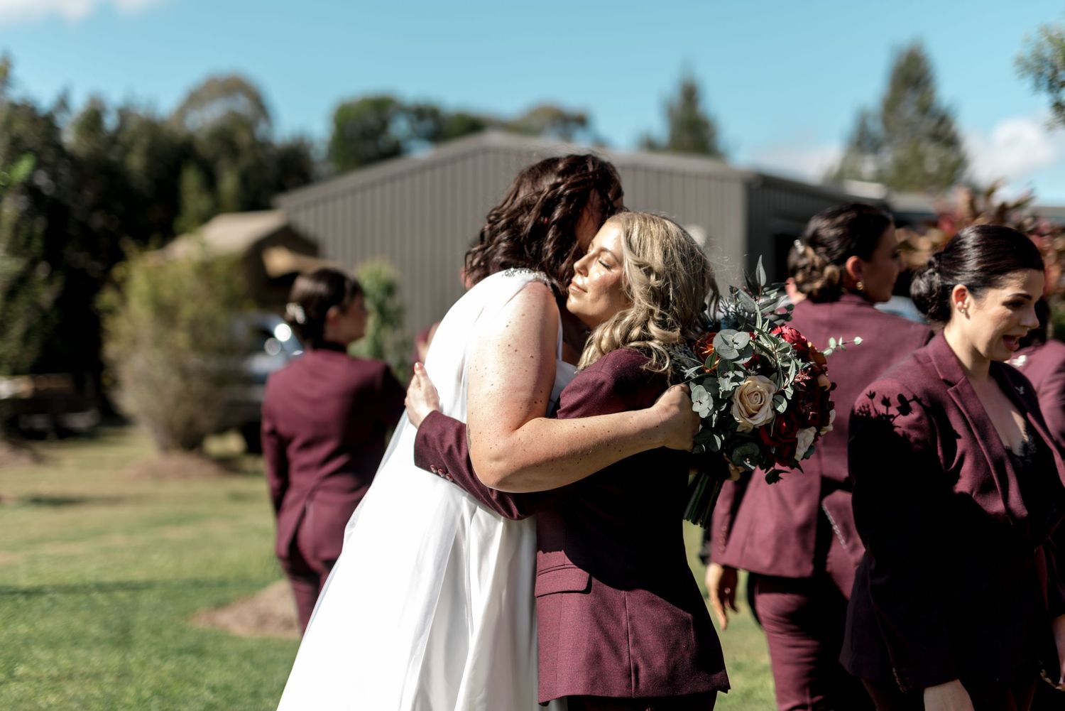 Bride shares emotional hug with bridesmaid in burgundy suit after outdoor wedding ceremony.