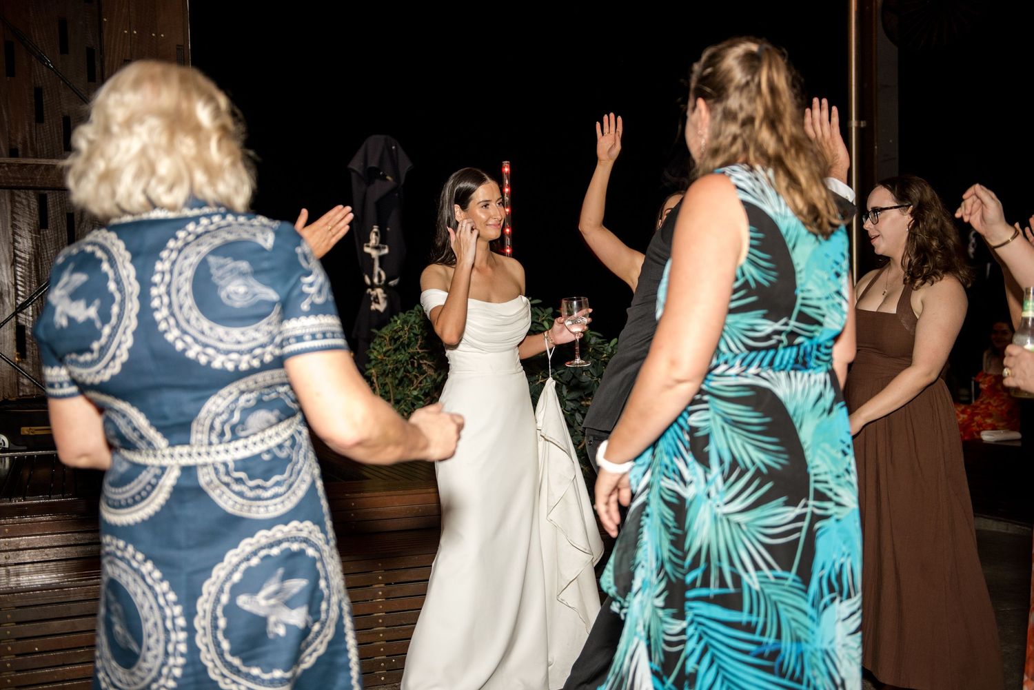 Bride dances and celebrates with female wedding guests during lively indoor reception dance floor moment.