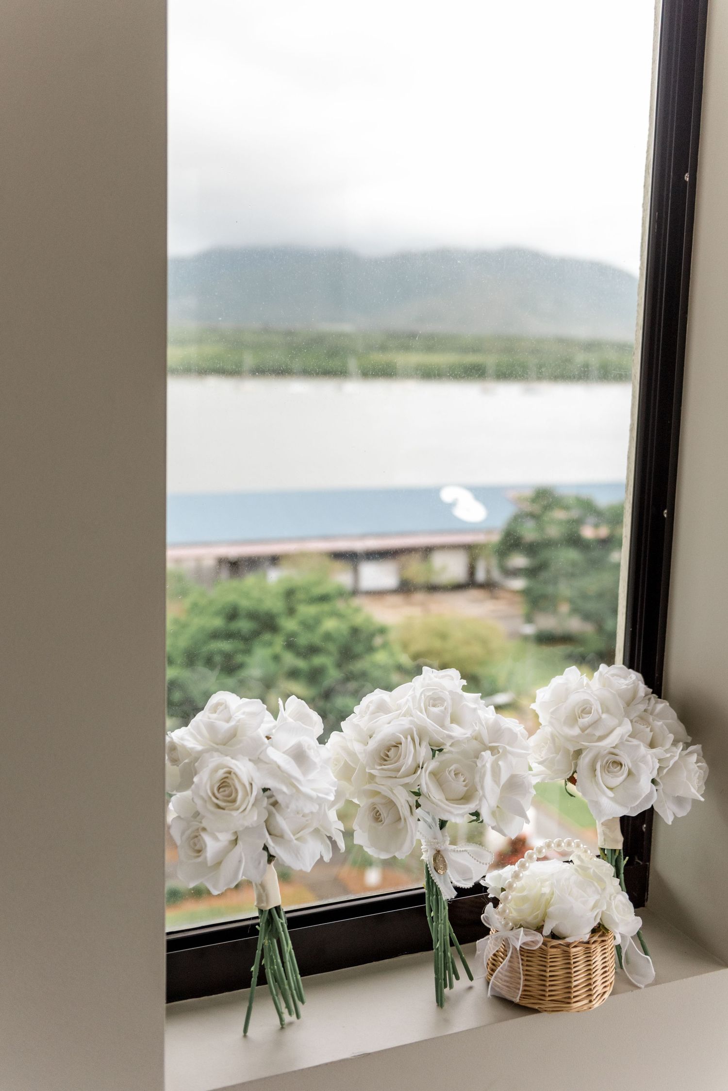 Three white rose bridal bouquets arranged on windowsill with scenic waterway and mountain views beyond.