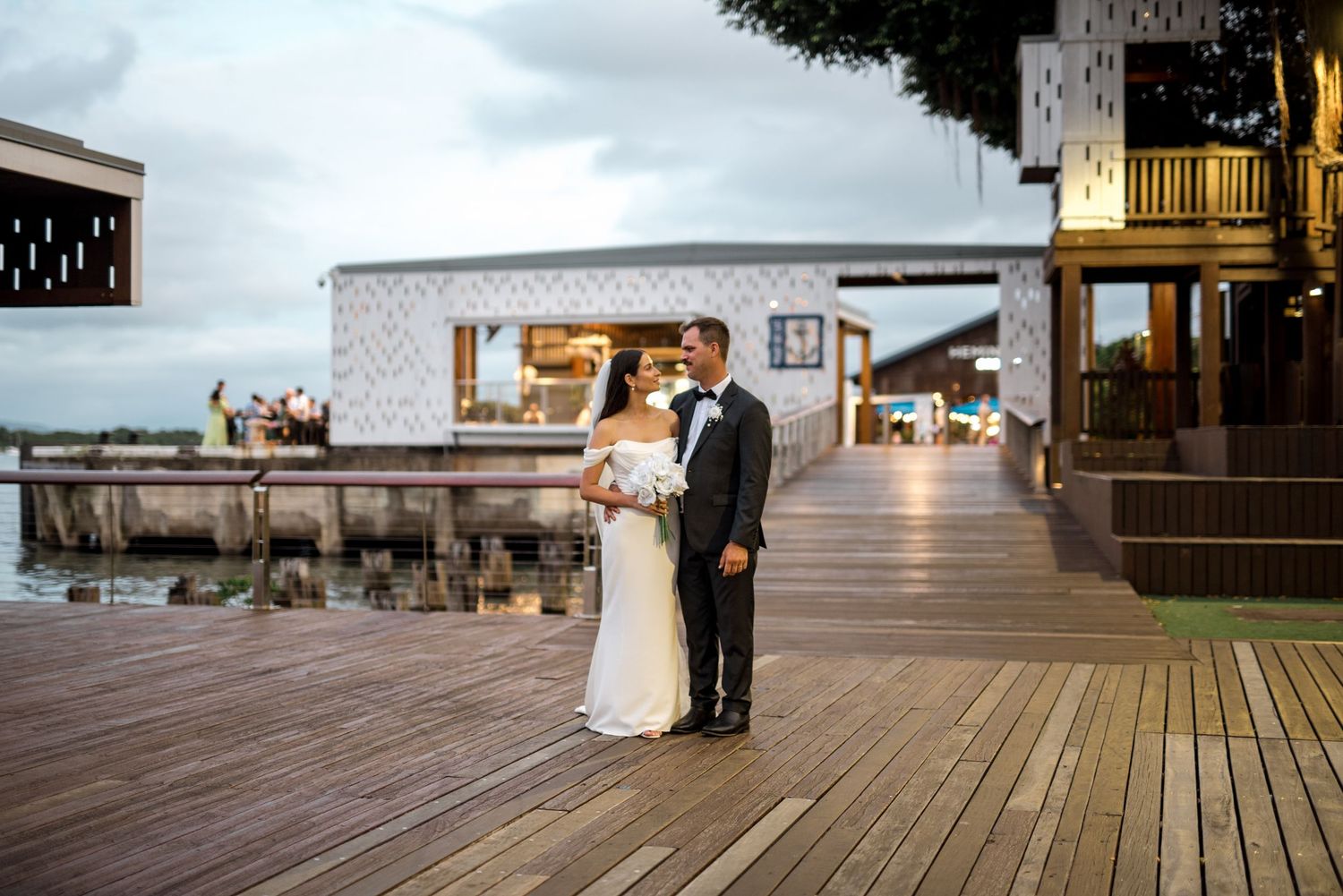 Bride and groom sharing a romantic moment on a wooden waterfront deck at dusk with venue lights glowing behind them.