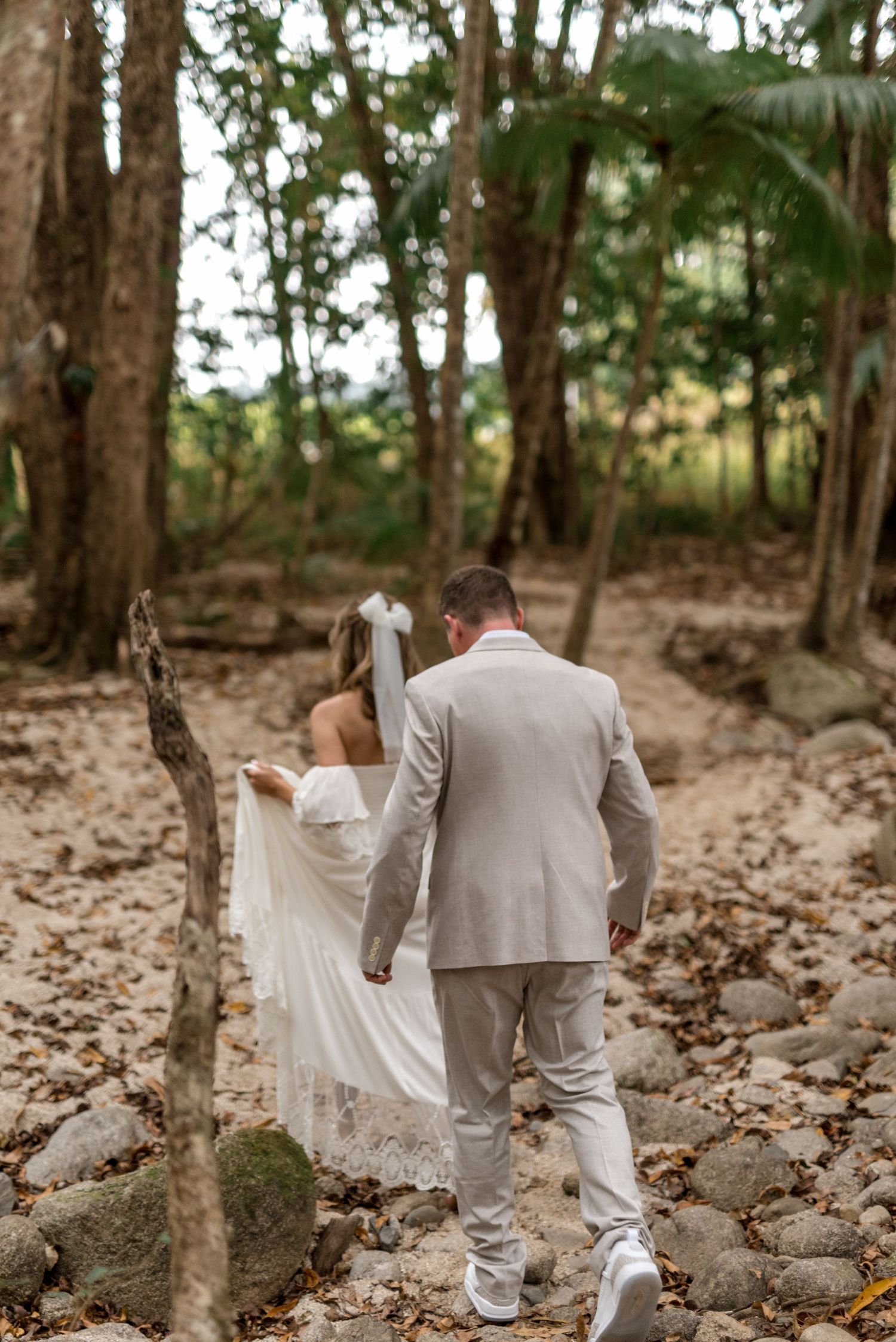 Bride and groom walking away through rocky forest path, bride in white lace gown with veil, groom in beige suit.