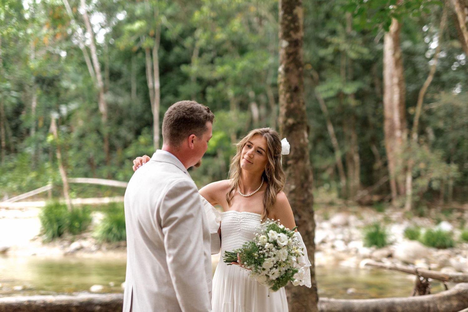 Groom tenderly holds bride's hand as she smiles holding a white bouquet in a forest setting.