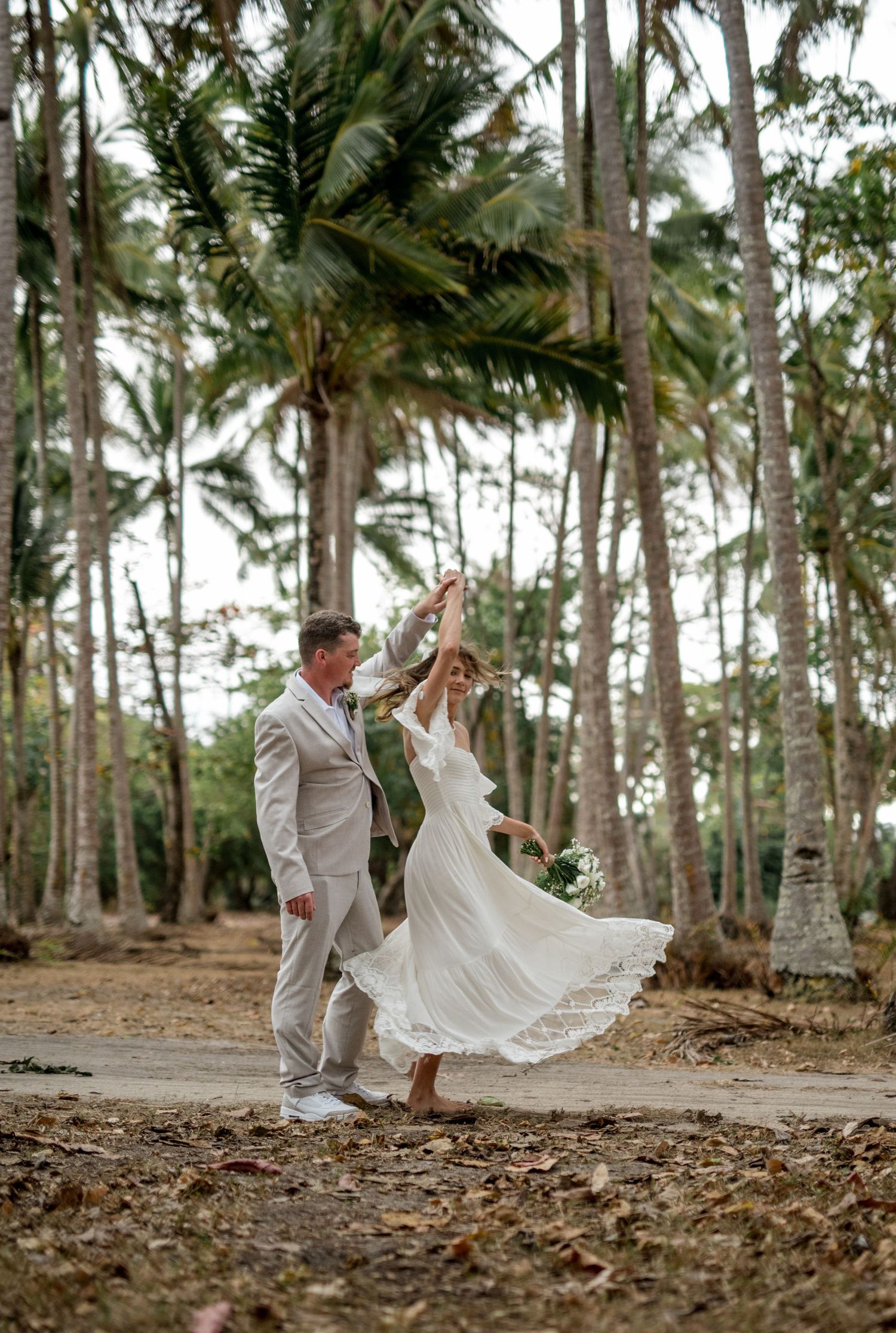 Bride twirls in white lace wedding dress while groom leads her through tropical palm tree forest.