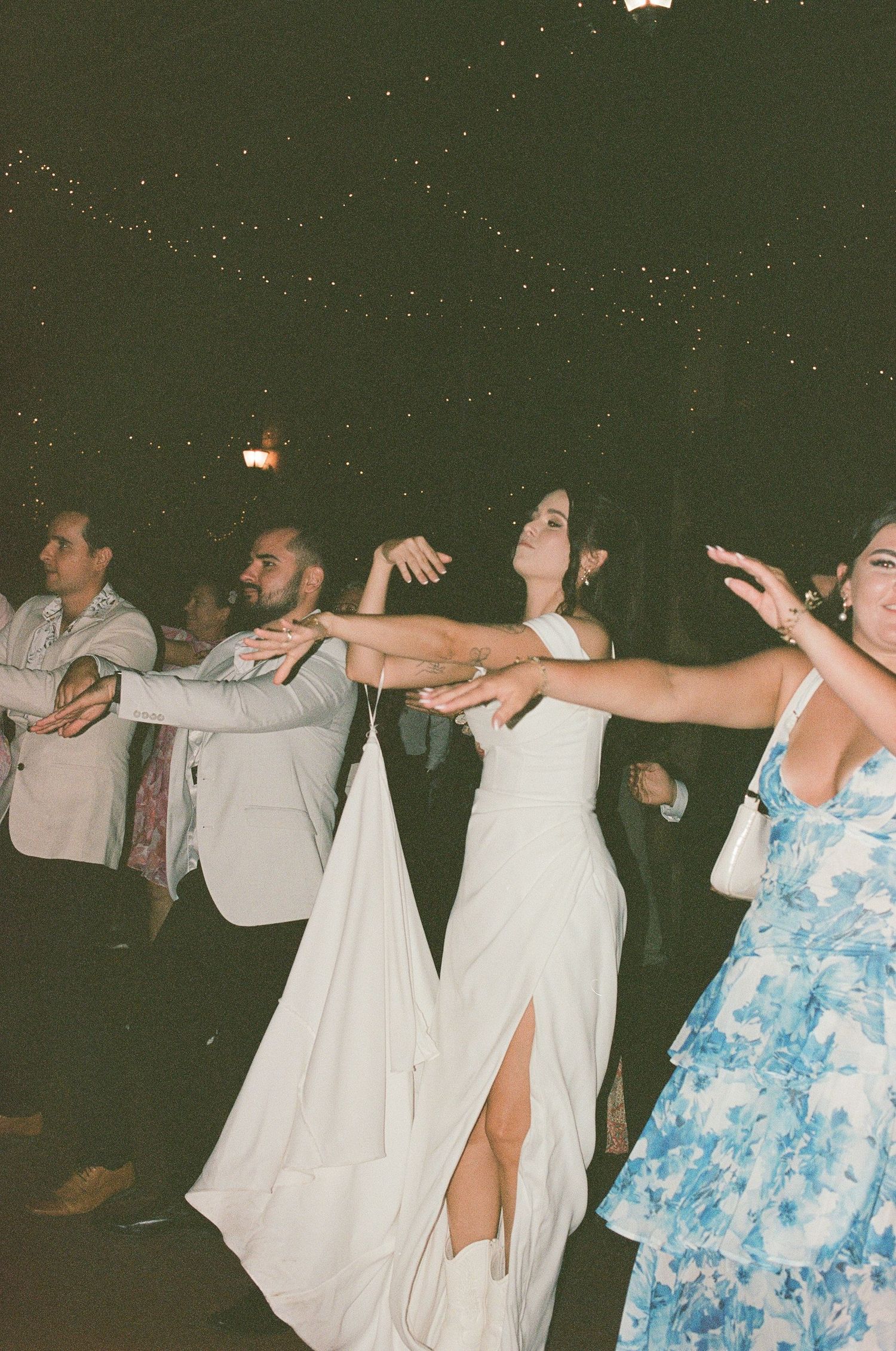 Bride in white slit gown dancing with arms outstretched at outdoor wedding reception.
