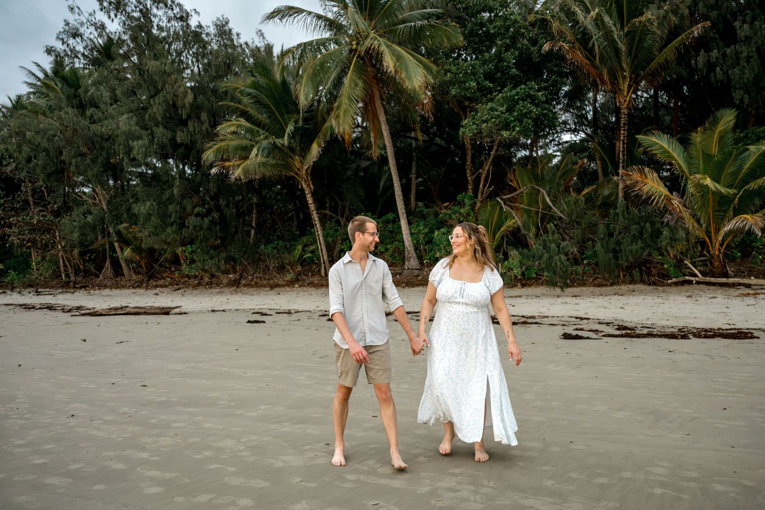 Happy couple holding hands and strolling along sandy beach lined with lush tropical palm trees at Four Mile Beach, Port Douglas.