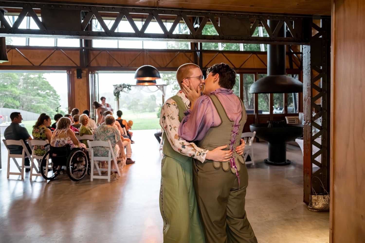 Two brides share romantic kiss after wedding ceremony in rustic indoor venue with guests seated behind them.