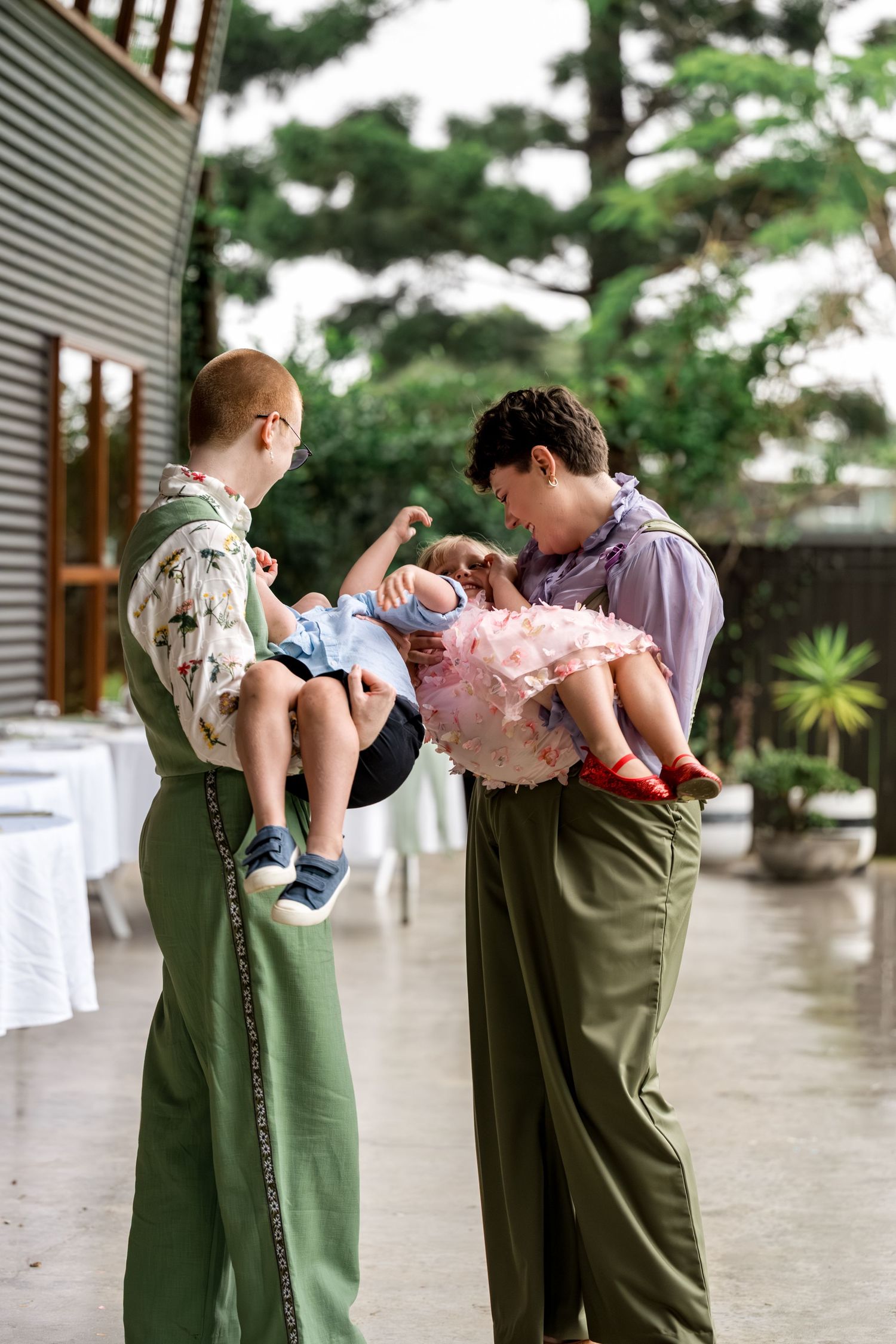Two people in matching green outfits each hold a baby at a wedding venue.