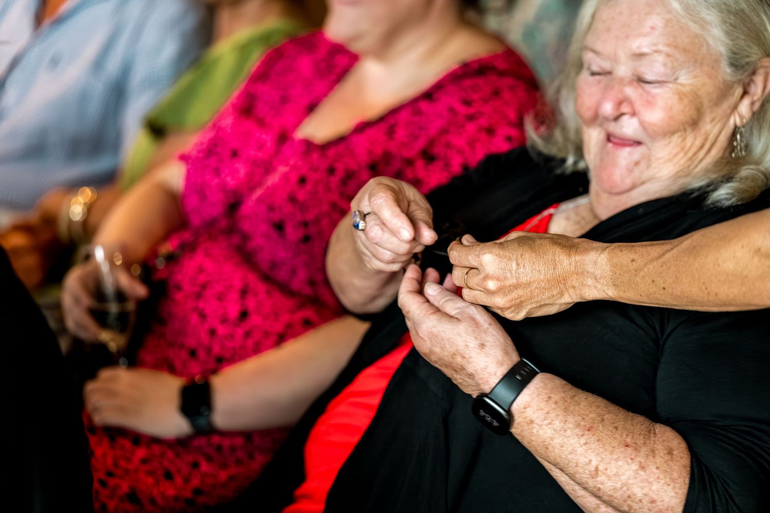 An elderly woman in black smiles while examining jewelry at a wedding celebration event.