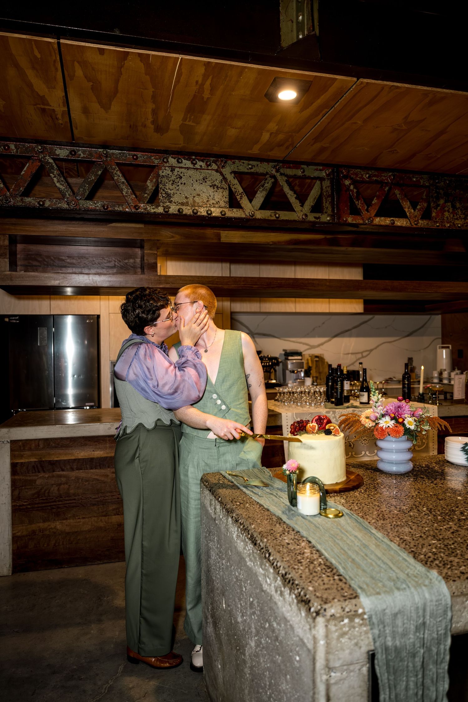 Newlyweds share a kiss while cutting their wedding cake at a rustic venue.