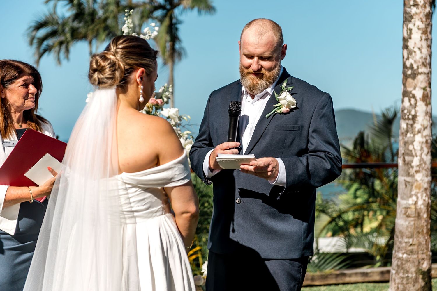 Groom reads vows holding a ring during outdoor wedding ceremony with officiant and tropical scenery behind.