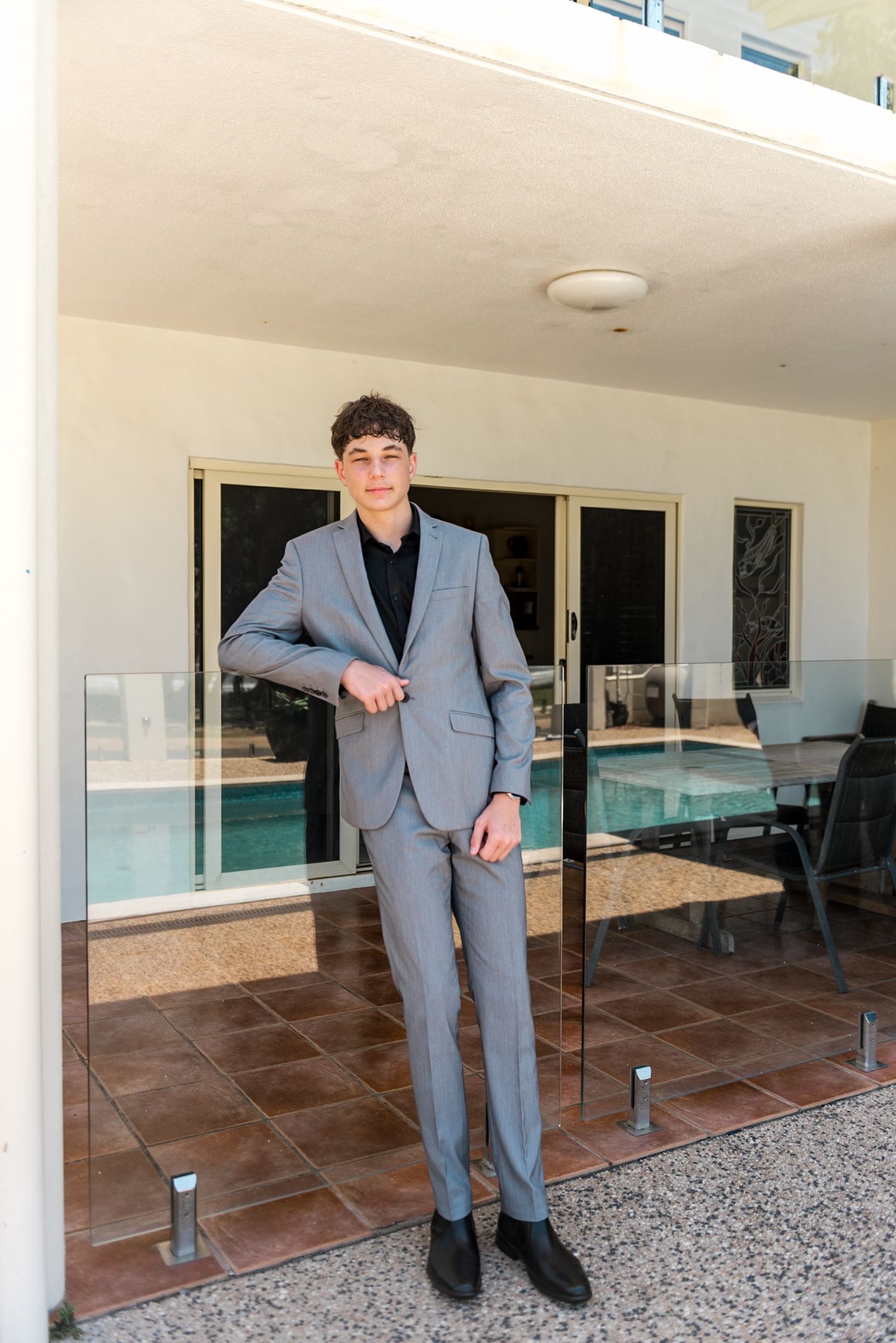 Young man in a grey suit posing confidently on a tiled patio near glass railings.