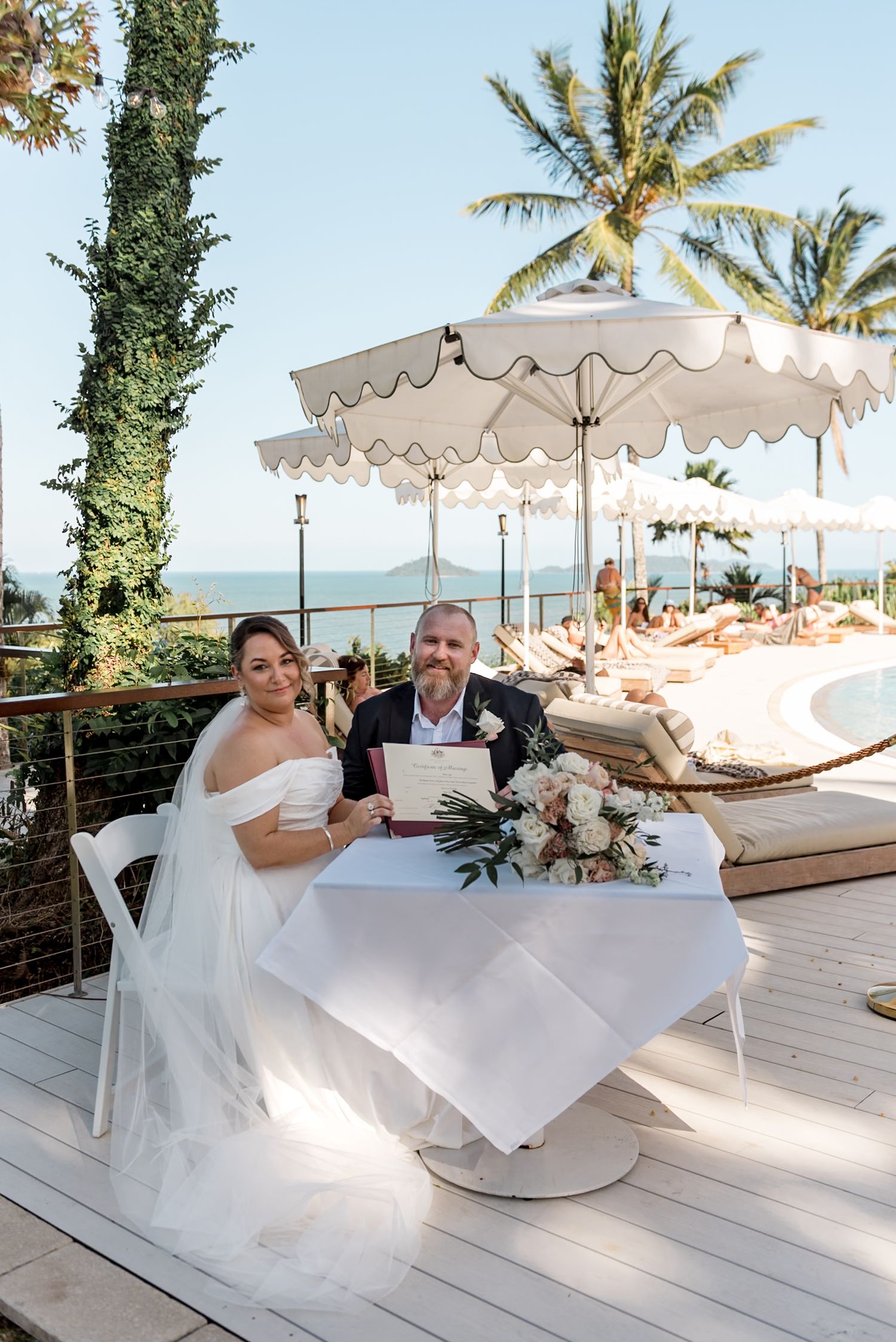 Newlyweds proudly displaying marriage certificate at outdoor tropical venue with ocean backdrop.