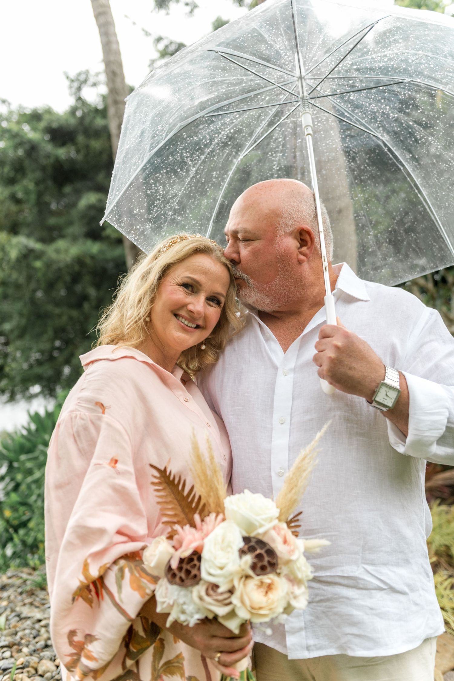 Couple sharing a romantic kiss under a clear bubble umbrella holding a bouquet of white and blush flowers.