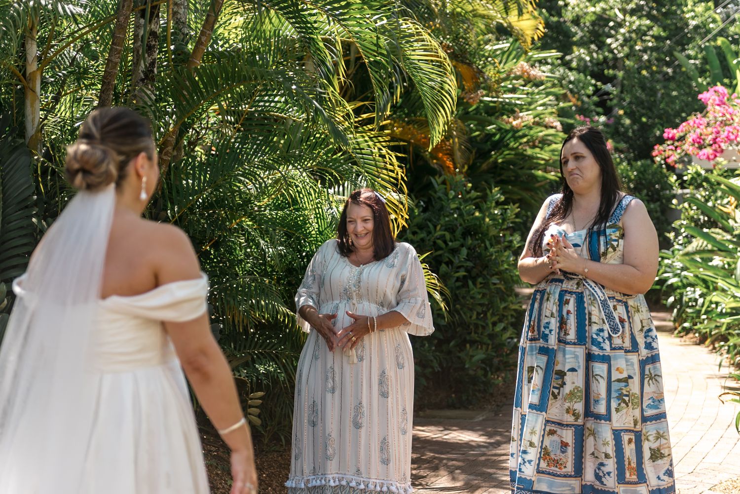 Bride in white gown and veil greeted by two women in tropical garden with pink flowers.