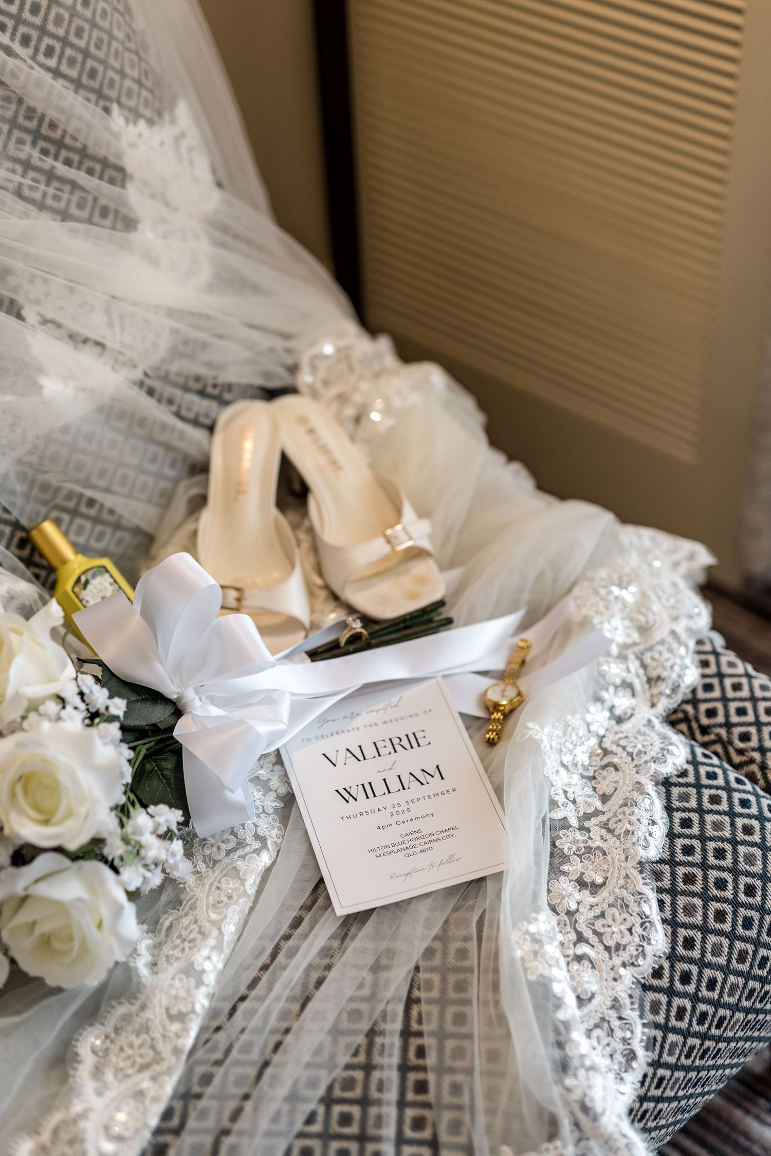 Bridal details flat lay with white sandals, roses, Valerie and William wedding invitation, and perfume on lace veil.