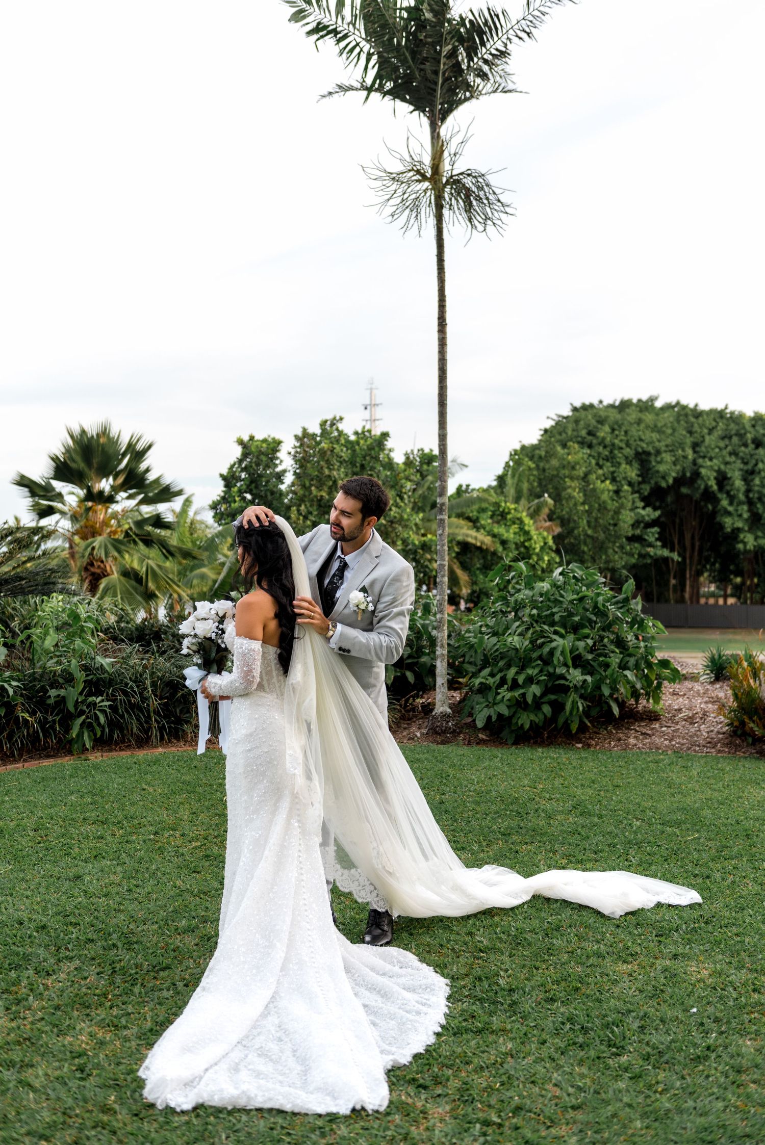 Groom in grey suit adjusting bride's long veil in lush tropical garden setting.