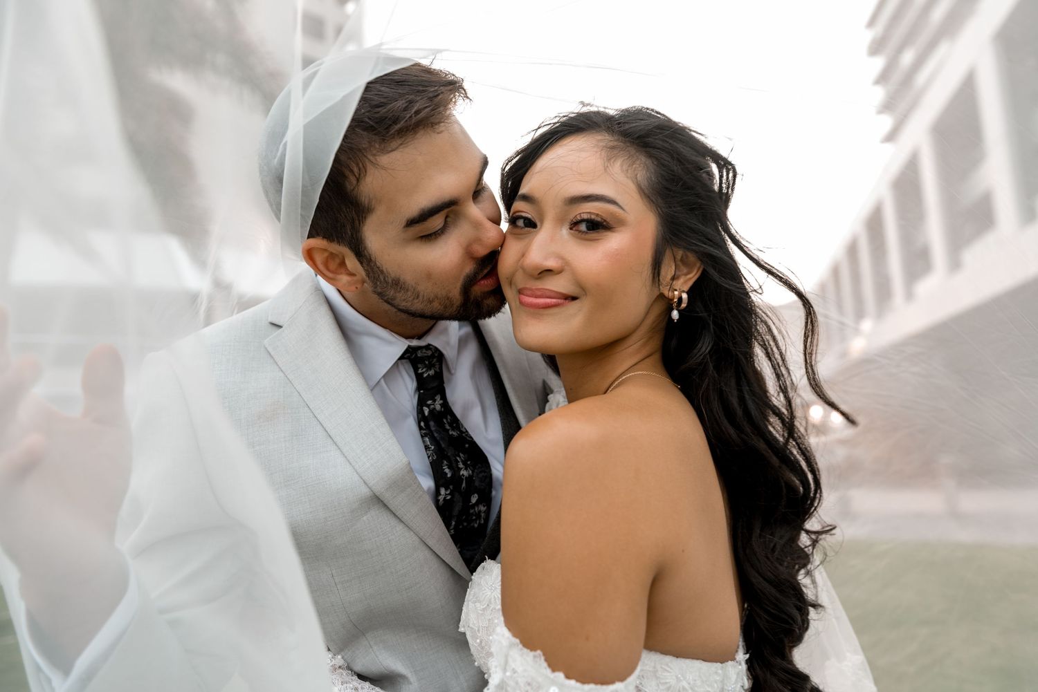 Groom kisses bride's cheek tenderly under a white veil as she smiles beautifully at the camera.