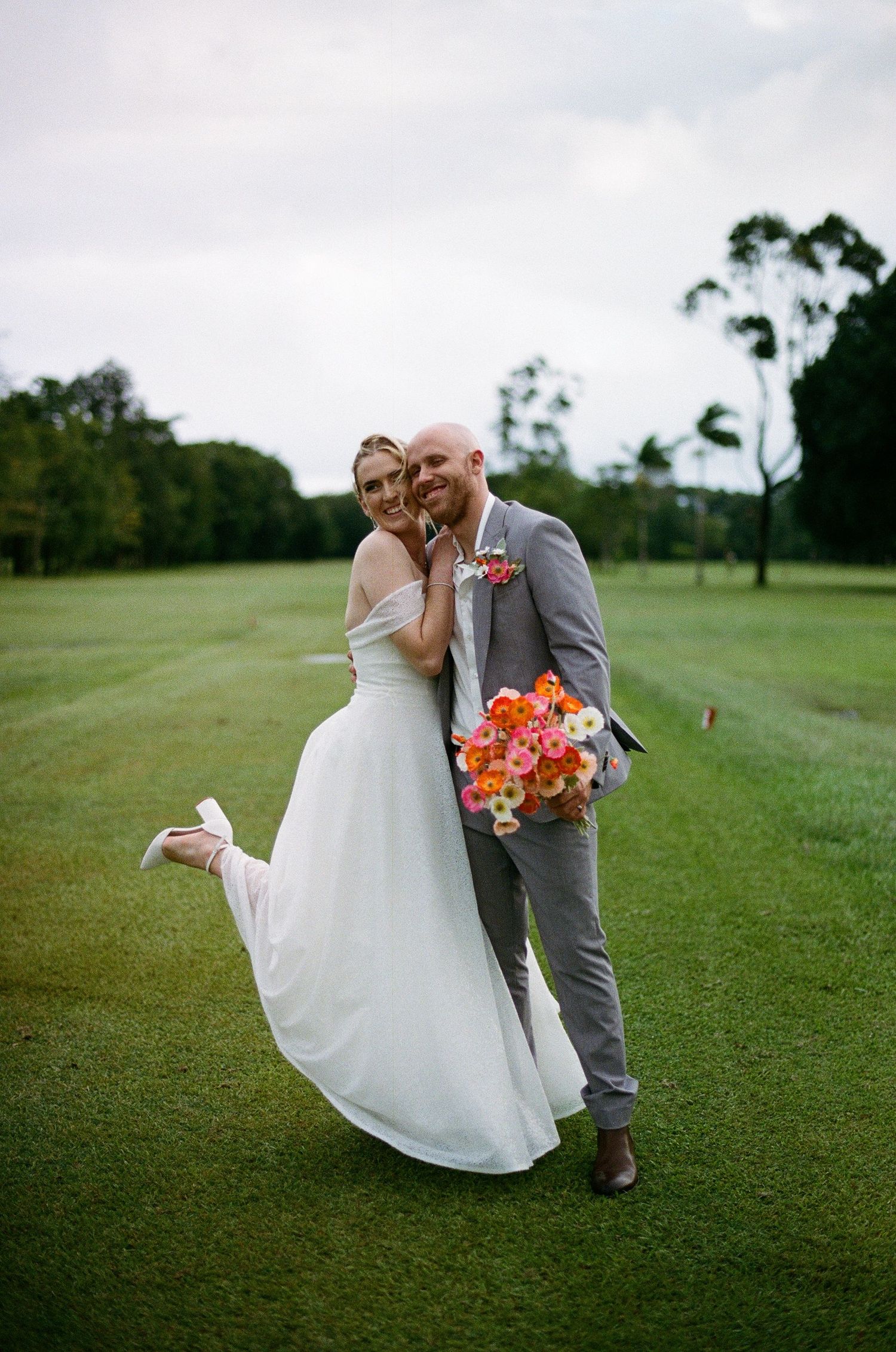 Bride and groom posing joyfully on a lush green golf course, groom holding a vibrant pink and orange bouquet.