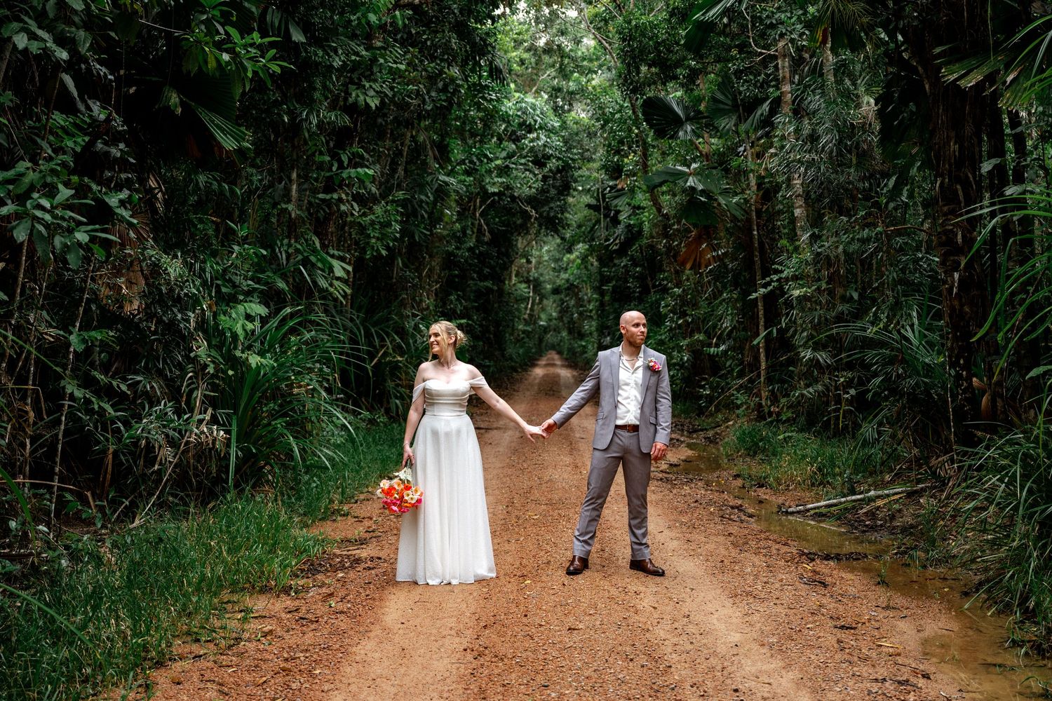 Bride and groom hold hands apart on a lush tropical dirt road, bride holding bright orange bouquet.