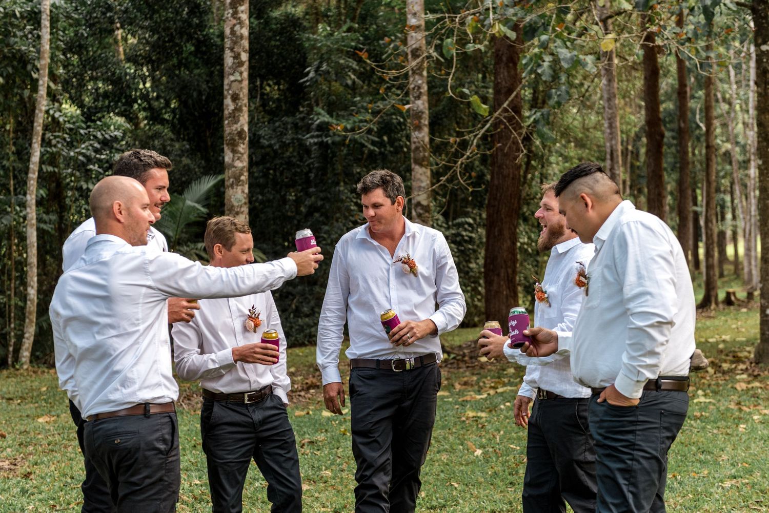 Five groomsmen in white shirts laugh and celebrate with drinks in a lush forest wedding setting.