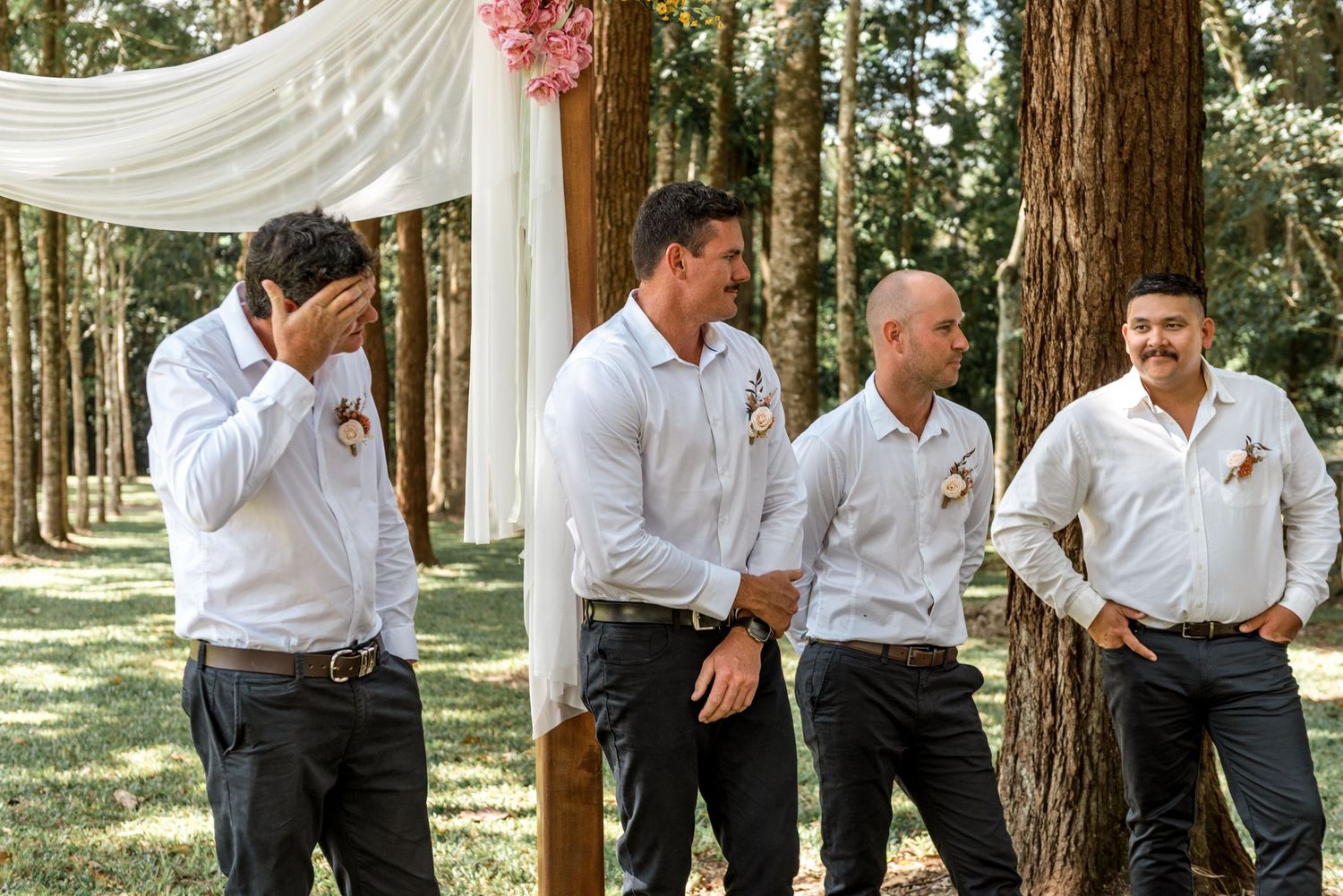 Groomsmen in white shirts laugh together beside a floral wedding arch in a pine forest.